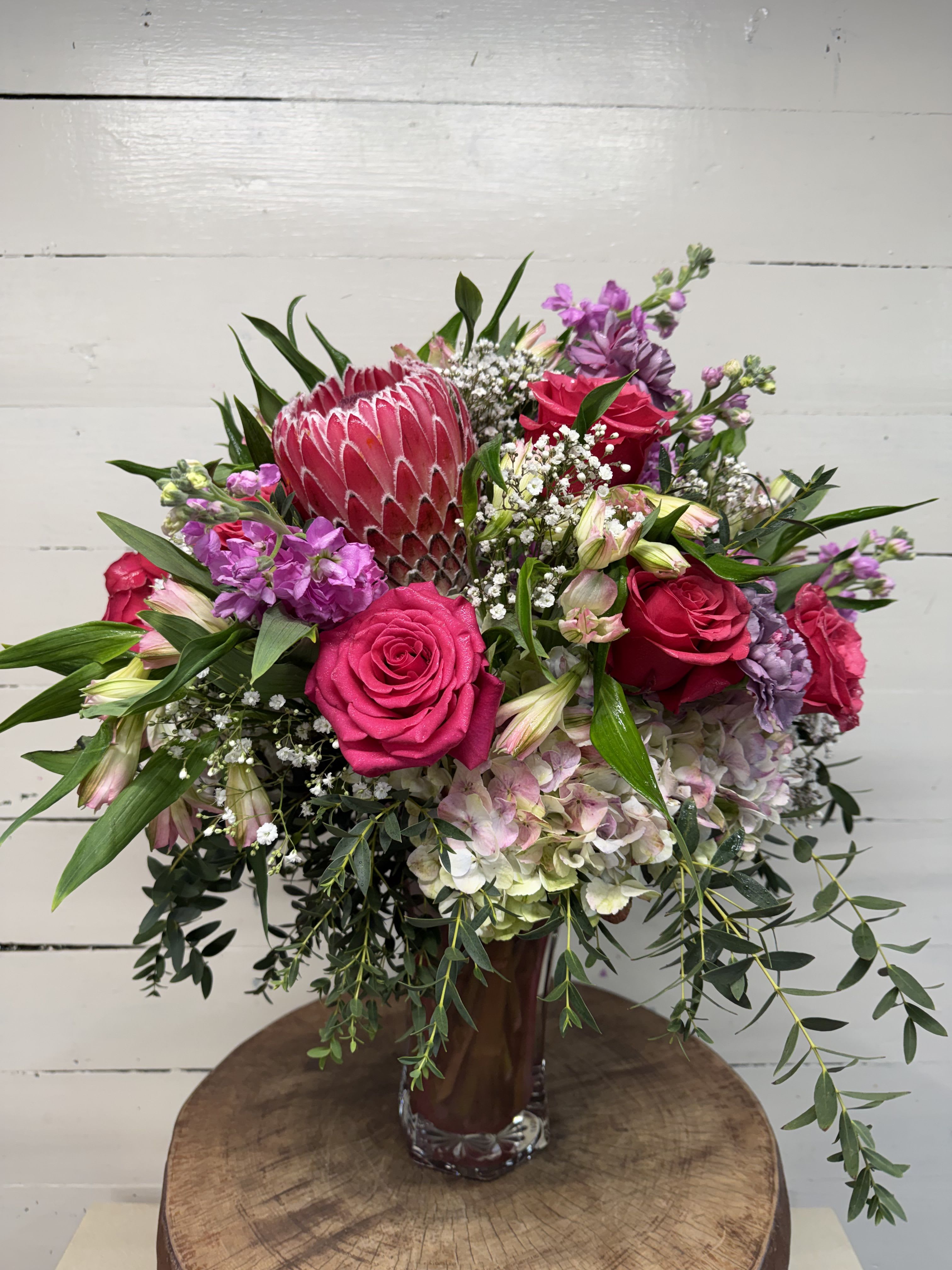 WCF Luxe Collection - One in a Million - Surprise your special one with this gorgeous arrangement and show them that they are truly one in a million! Let this grand bouquet in the perfect blend of pinks and purples show them that they are truly seen, heard, loved and valued.   Includes one dozen long stem hot pink roses, pink hydrangeas, stock, alstroemeria, carnations and featuring a gorgeous pink protea and accented with fresh greens. Delivered in a frosted pink glass vase. Approximately 21 3/4" W x 25" H  Please note that orders must be placed a least 48 hours prior to fulfillment date in order to procure the freshest and highest quality blooms. 