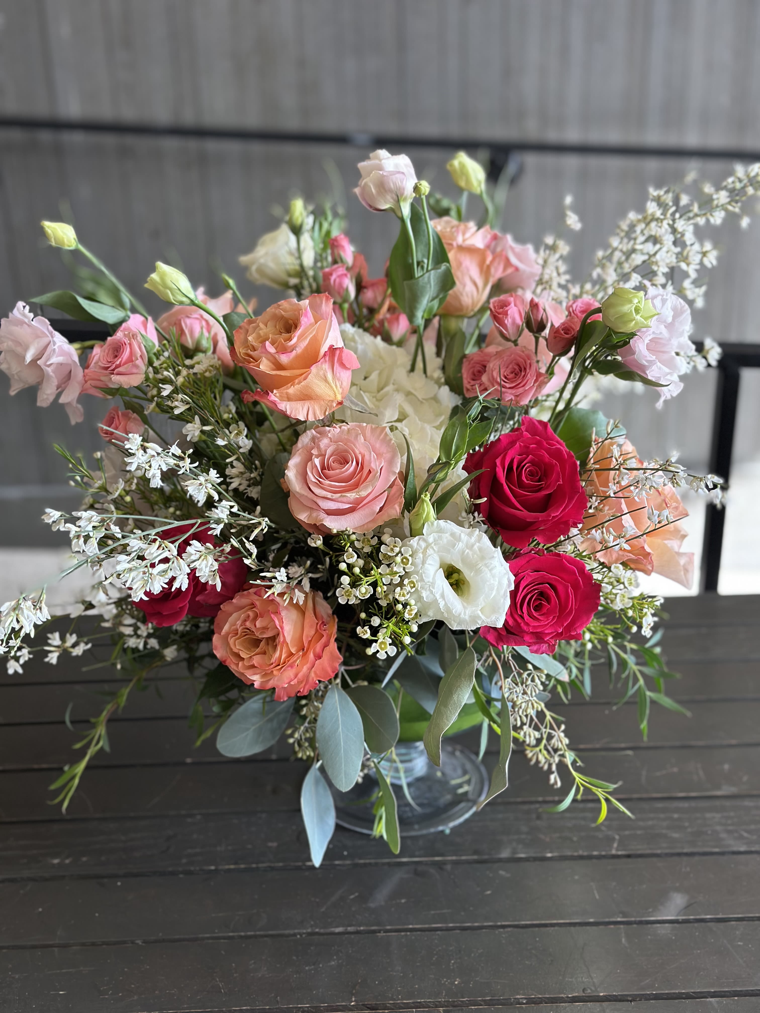 Pink Lizzy - A round glass pedestal filled with pink, white lisianthus, roses, hydrangeas, white ginestra, and greenery. 