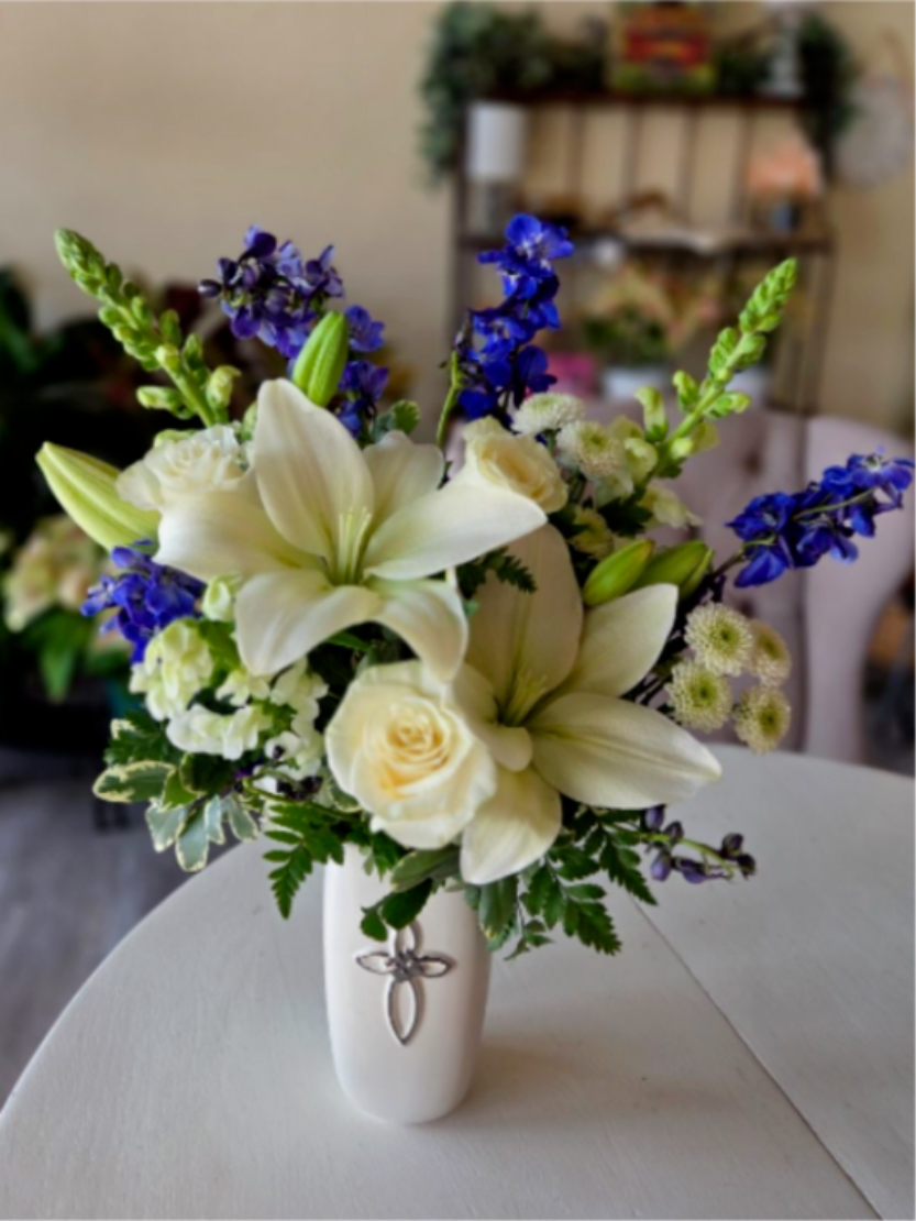Hopeful Blessings - Featuring pure white lilies and roses, symbolizing faith, renewal, and everlasting love. Accented with deep blue delphinium and gentle greenery, it is thoughtfully designed in a white ceramic vase with a cross, making it a meaningful expression of prayer, comfort, and hope during times of remembrance or spiritual reflection.