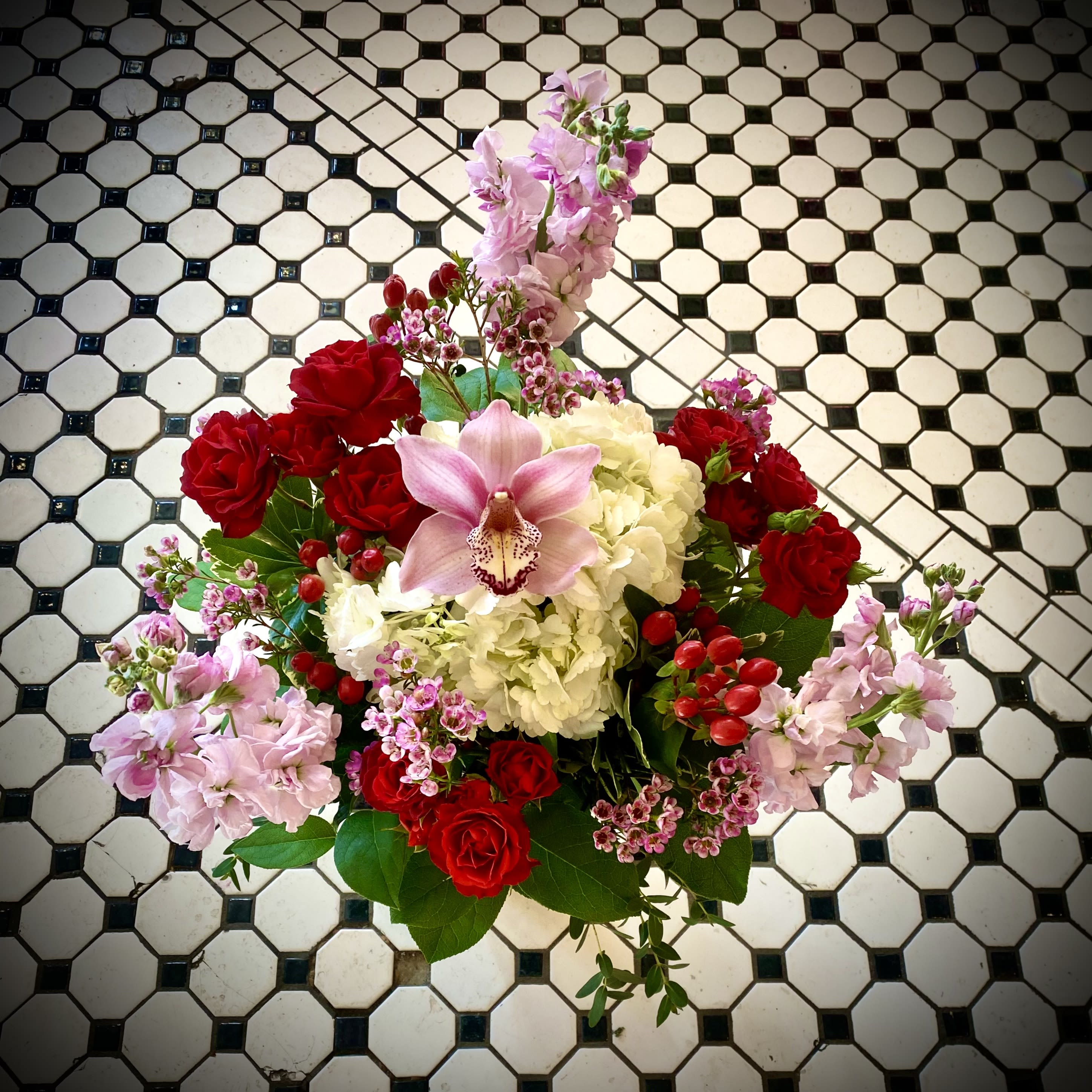 Cupid's Kiss - Romantic compact arrangement in shades of Pink, White and Red. Includes a Cymbidium orchid, White Hydrangea, Red Spray Roses, and berries designed in a beautiful red keepsake vase. Approximately 15 inches by 12 inches.