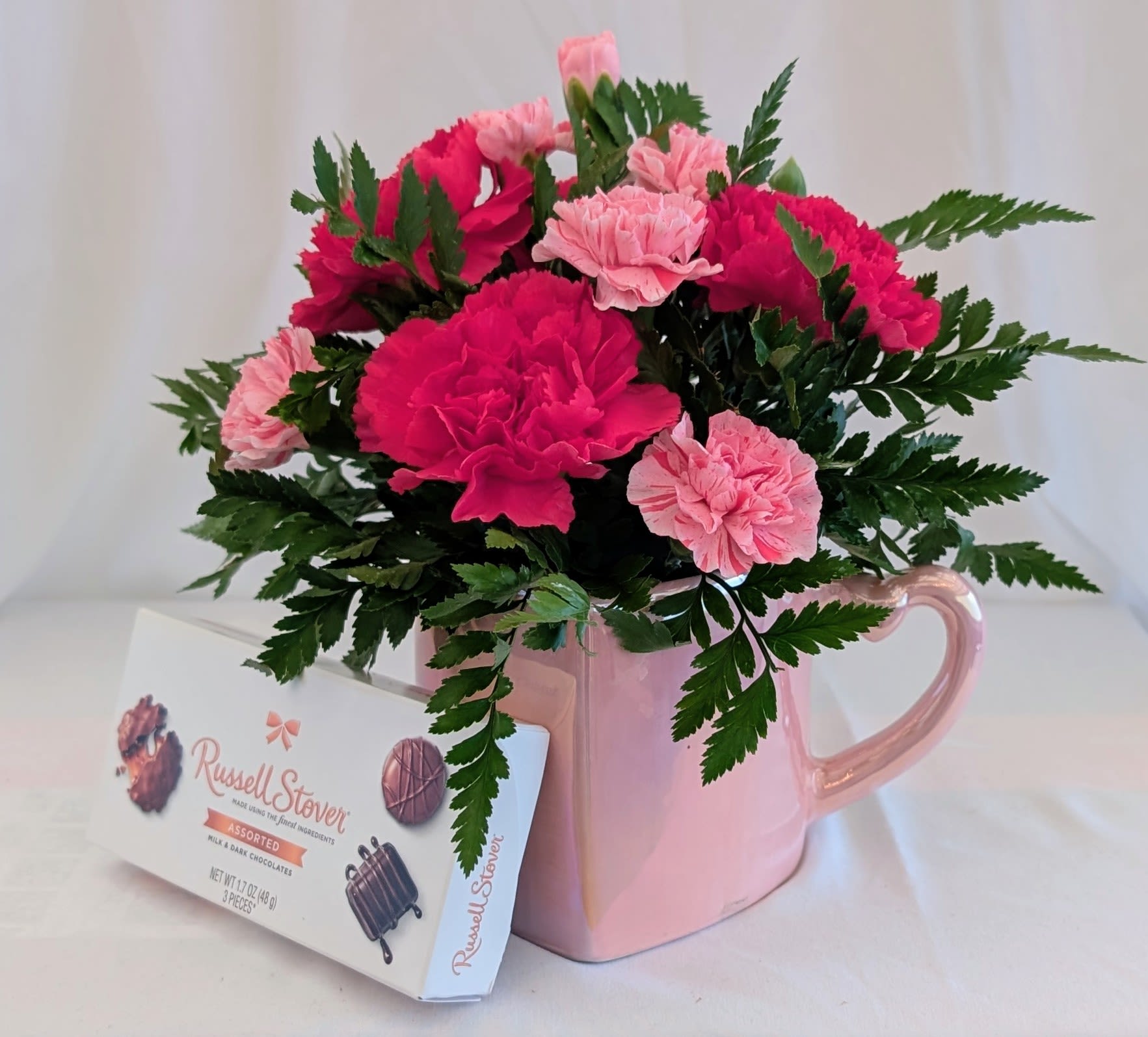 Pink Petals &amp; Chocolates - A charming burst of pink perfection! This sweet arrangement features fresh pink carnations nestled in a reusable heart-shaped ceramic mug (pink or white) accented with lush greenery for a full, cheerful look. Paired with a mini-box of Russell Stover® assorted chocolates, it’s the perfect way to say “thinking of you,” “thank you,” or “just because.” Flower shades may vary slightly based on availability, but the beauty and value remain the same. WHITE MUGS ALSO AVAILABLE.