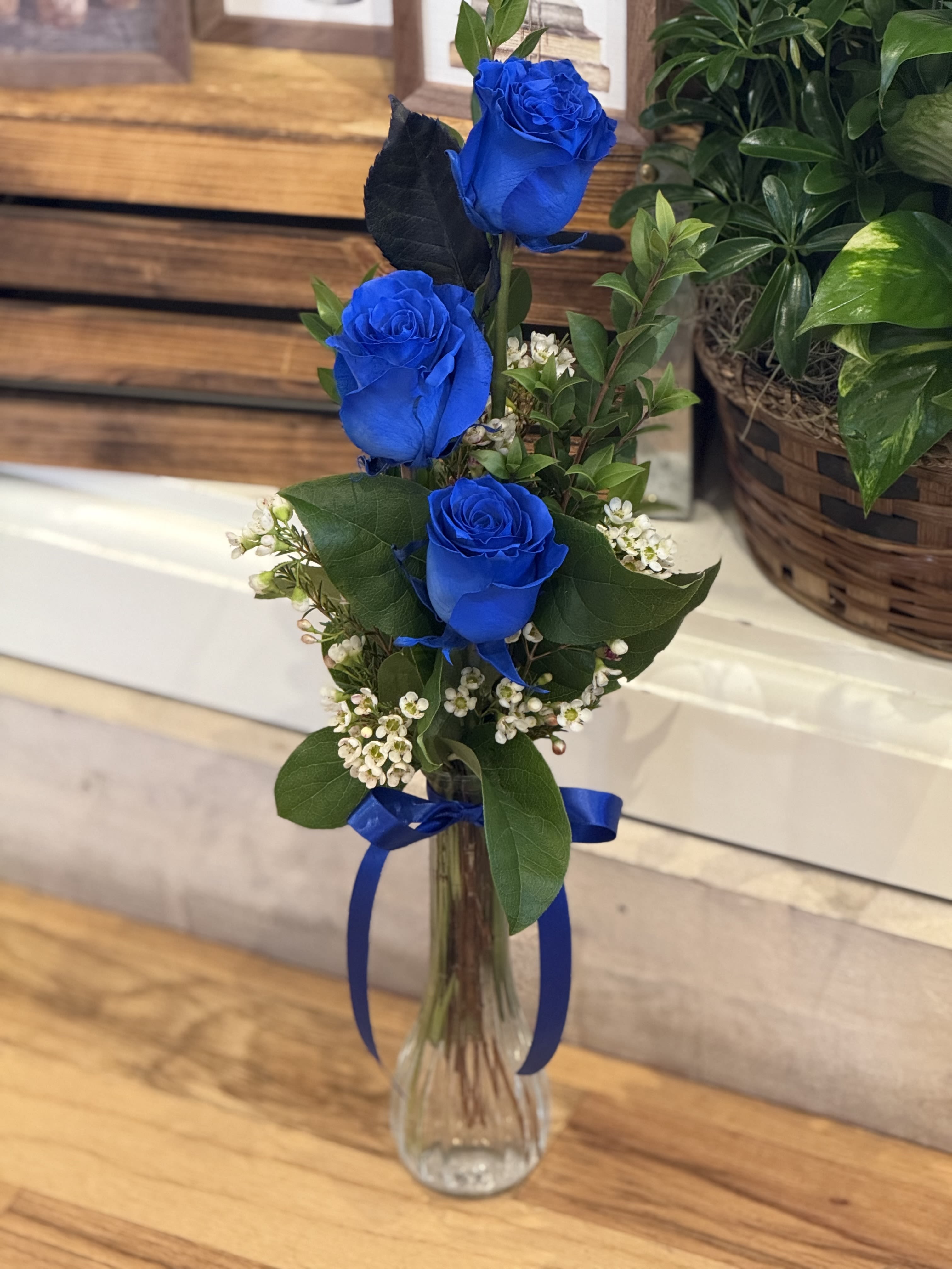 Three Blue Roses - Three blue roses nestled in assorted greenery in a bud vase. Finished off with wax flower and coordinated ribbon. 