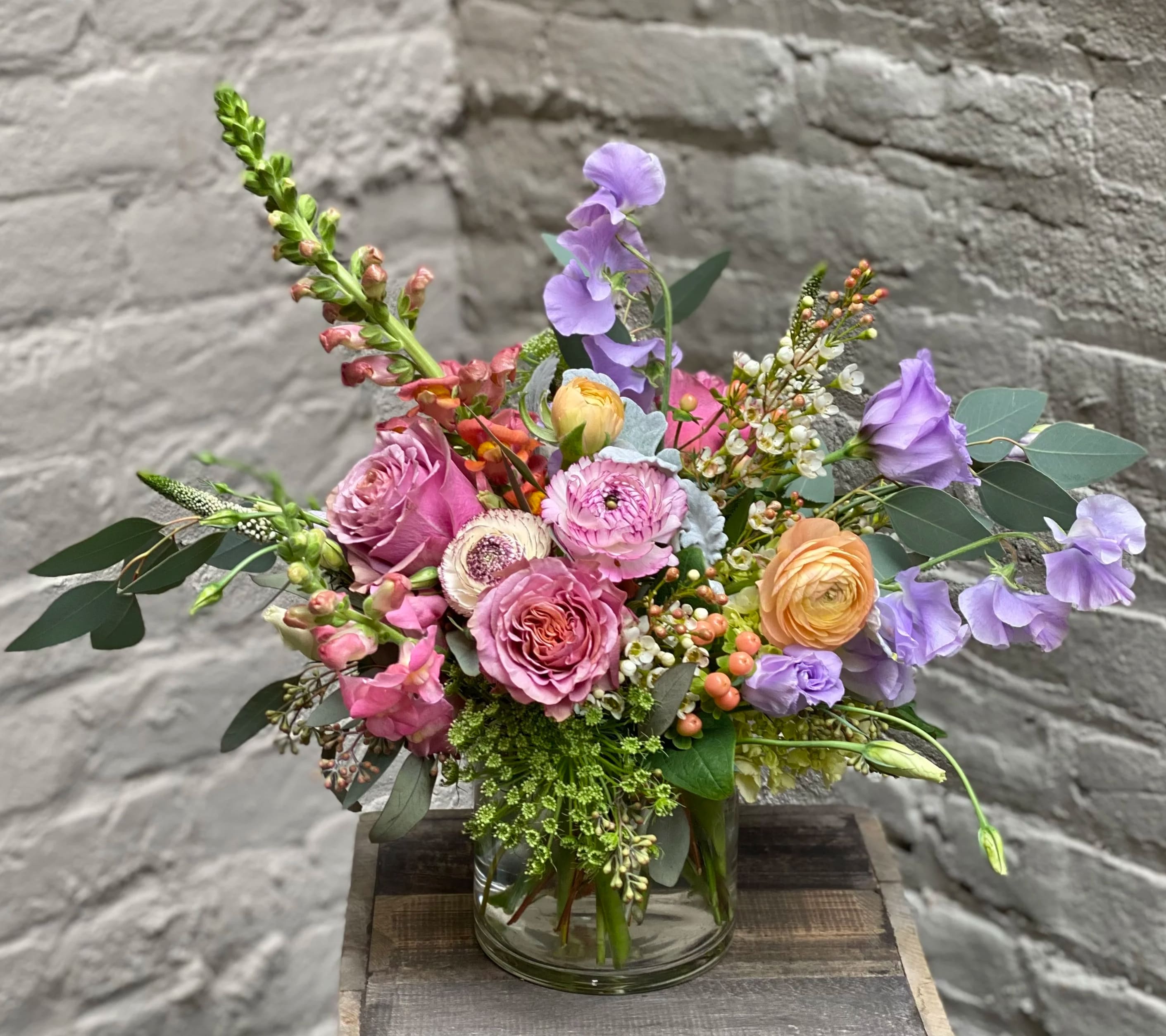 Ostara - Beautiful purple, pink and peach blooms come together to make this harmonious display of springtime flowers, including sweet peas, ranunculus and snapdragons.  Named for the ancient Germanic goddess of spring, this glass vased arrangement is the perfect way to celebrate a season of life and new growth.  Arrangement measures approximately 12x12".