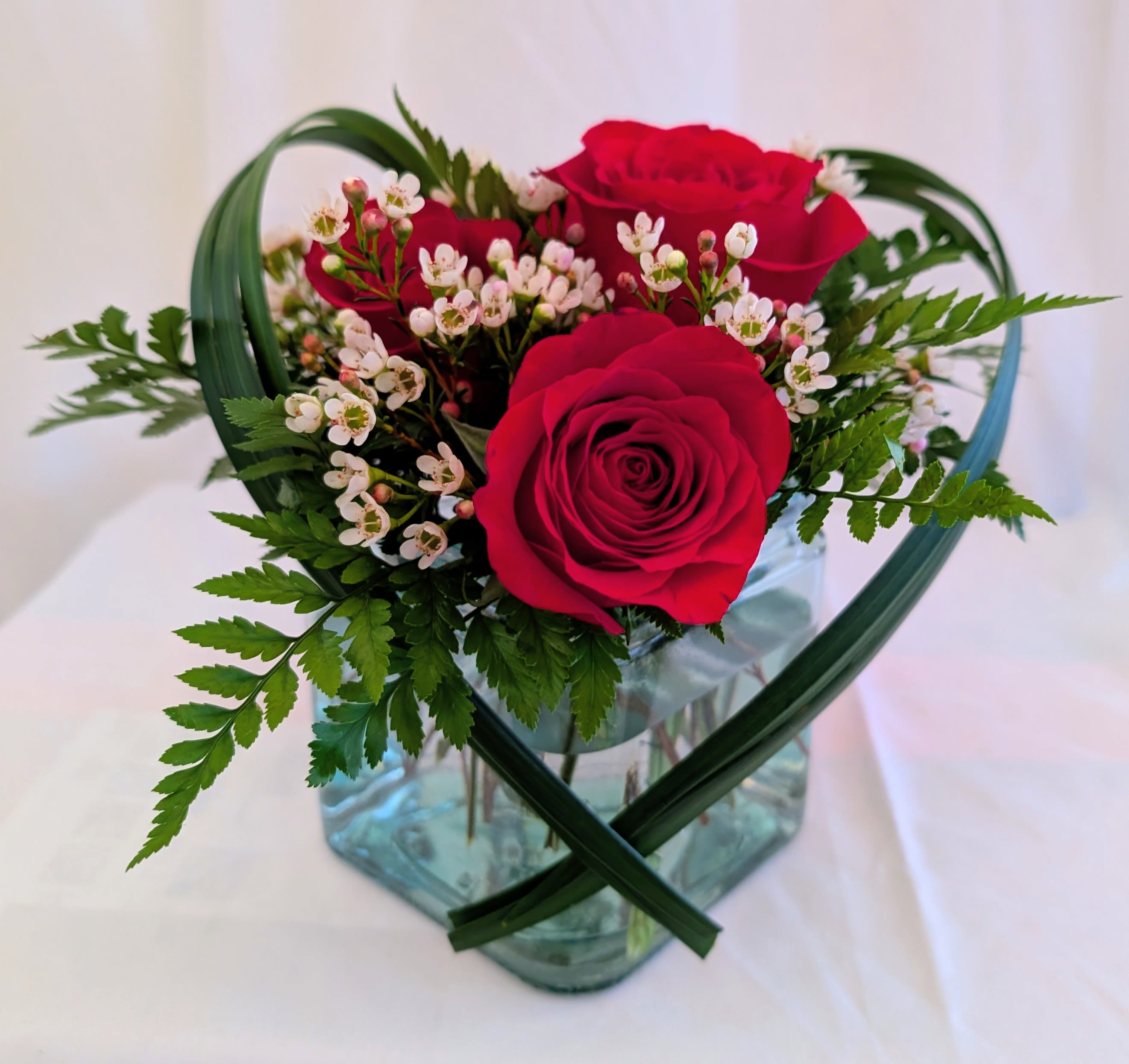 Simply Yours - Elegant and expressive, this arrangement features vibrant red roses surrounded by soft white accent blooms and fresh greens, designed in a clear glass cube with a heart-inspired foliage flourish. A beautiful way to say “I love you” without saying a word. Some substitutions may occur while preserving overall look and feel.