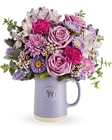 Teleflora's Sweetest Flutter Bouquet - Gift mom the artisanal charm she loves with the Sweetest Flutter glazed ceramic pitcher, adorned with embossed butterfly detail and a natural base, perfect for year-round use and sure to add a touch of elegance to any table setting. Surprise her with the Sweetest Flutter bouquet-lavender roses, alstroemeria, fuchsia carnations, matsumoto asters, cushion spray chrysanthemums, sinuata statice, pink waxflower, dusty miller, and spiral eucalyptus-in the food-safe glazed ceramic pitcher, featuring embossed butterfly detail, a cherished gift for the artisanal-loving mom.