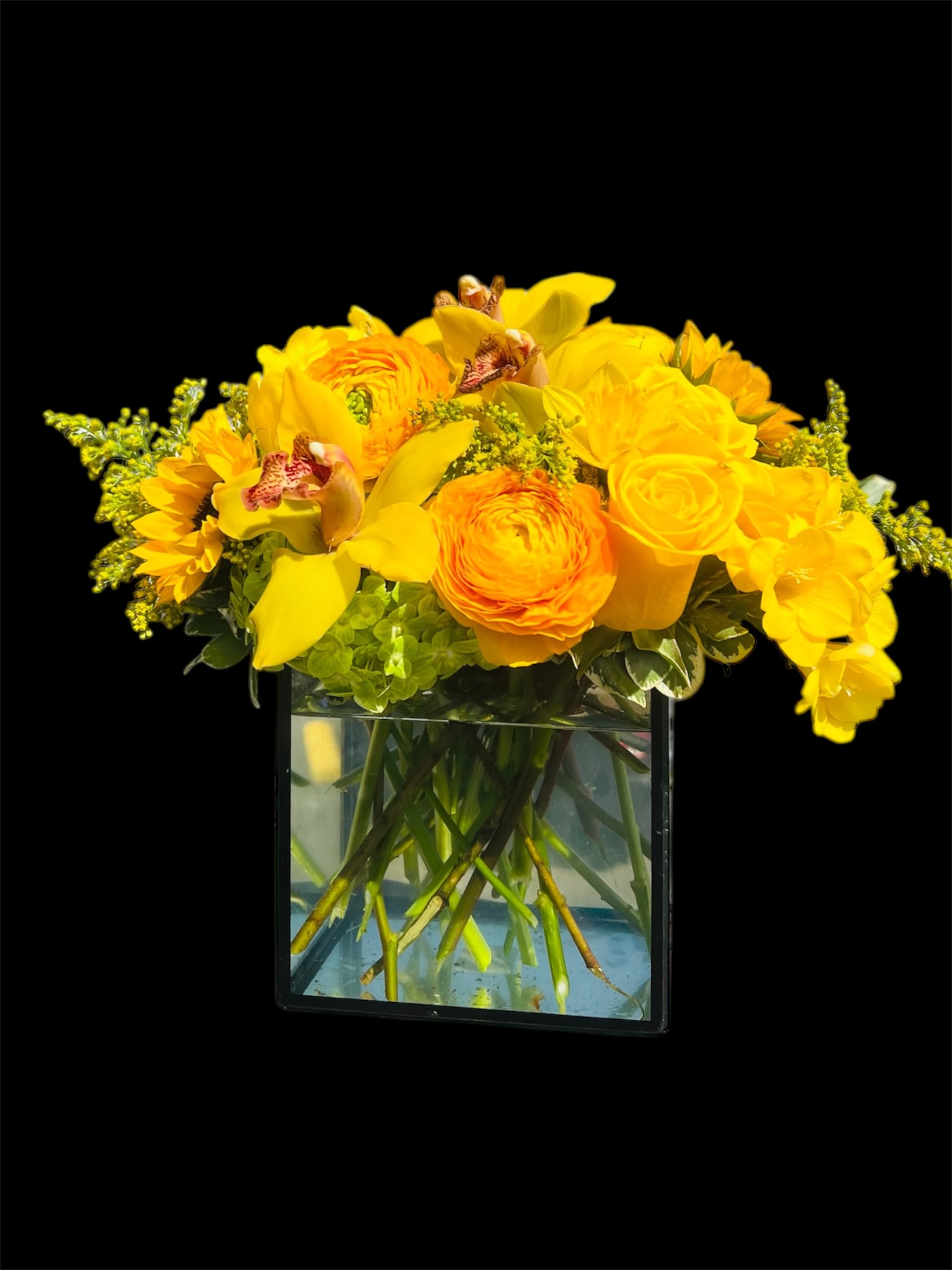 Bloom and glow  - A cheerful and vibrant arrangement showcasing the sunny shades of yellow blooms, including delicate ranunculus, elegant cymbidiums, classic roses, bright daffodils, graceful tulips, and bold sunflowers. These radiant flowers are accented by soft fillers that add texture and balance, creating a lively and harmonious display. Presented in a sleek cube glass vase, the arrangement radiates warmth and positivity, making it a perfect centerpiece to brighten any space.
