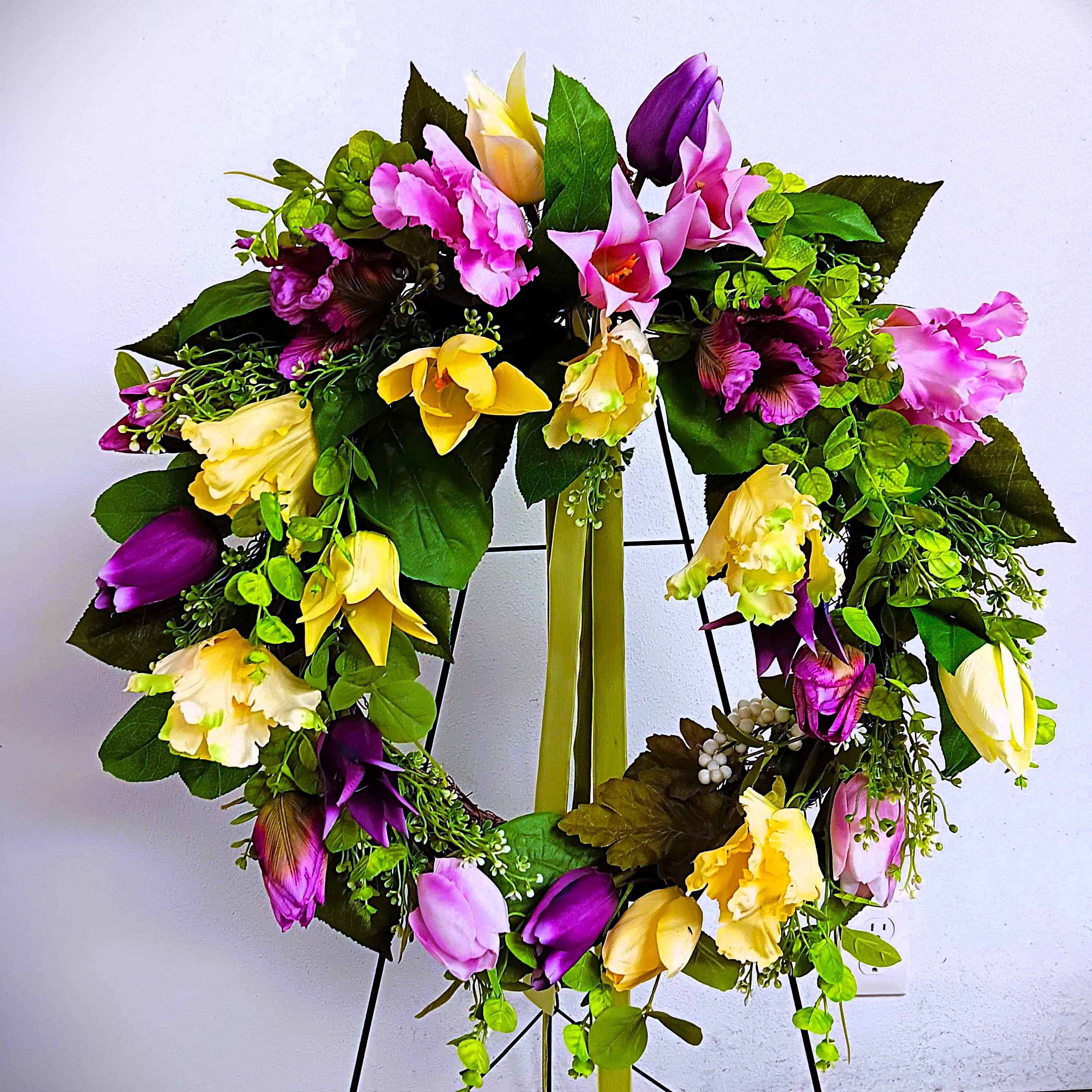 Lovely Lavender Easel Wreath - This artificial floral wreath of lilies, crocuses, and tulips in lavender and light yellow shades, is great to order for  Sympathy and Funeral services. Why silk flowers instead of fresh? Because this wreath will last longer, and left on the grave after services, it will stay for weeks and maybe months before fading away. Approx. 20" in diameter. We deliver!