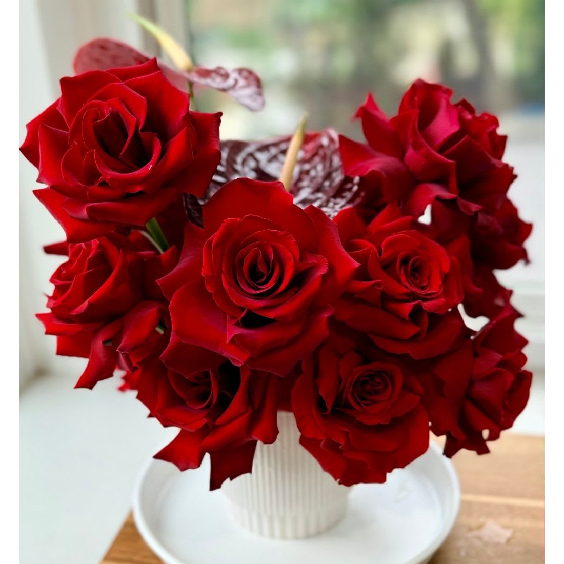 Crimson Romance - Crimson Romance – Luxe Hand-Reflexed Rose &amp; Anthurium Arrangement  This design offers a modern, architectural take on the classic red rose bouquet. We have paired the velvety texture of premium red roses with the glossy, sculptural lines of heart-shaped anthuriums to create a bold and sophisticated high-fashion aesthetic.  The delicate reflexing technique applied to each rose makes this arrangement truly unique. Our designers artfully fold back the outer petals by hand to create a voluminous, garden-style bloom. Please be assured that these roses are completely fresh; this specialized manipulation is purely stylistic, designed to maximize the flower's beauty and impact without compromising its quality or longevity.  Presented in a chic white ceramic vessel, Crimson Romance is an unforgettable statement perfect for anniversaries, Valentine’s Day, or any occasion that calls for a grand gesture.  Available Sizes:  • Standard – A refined presentation of 15 premium red roses accented with 2 anthuriums, elegant and impactful.  • Deluxe – A fuller, more indulgent design with additional roses and anthurium blooms for enhanced depth and drama.  • Premium – Our most opulent expression, featuring an abundant composition of roses and anthuriums for a striking, couture-style statement.  Care Tips:  • Display in a cool environment away from direct sunlight  • Refresh water daily and re-cut stems every 1–2 days  • With proper care, blooms will maintain beauty and presence for days to come