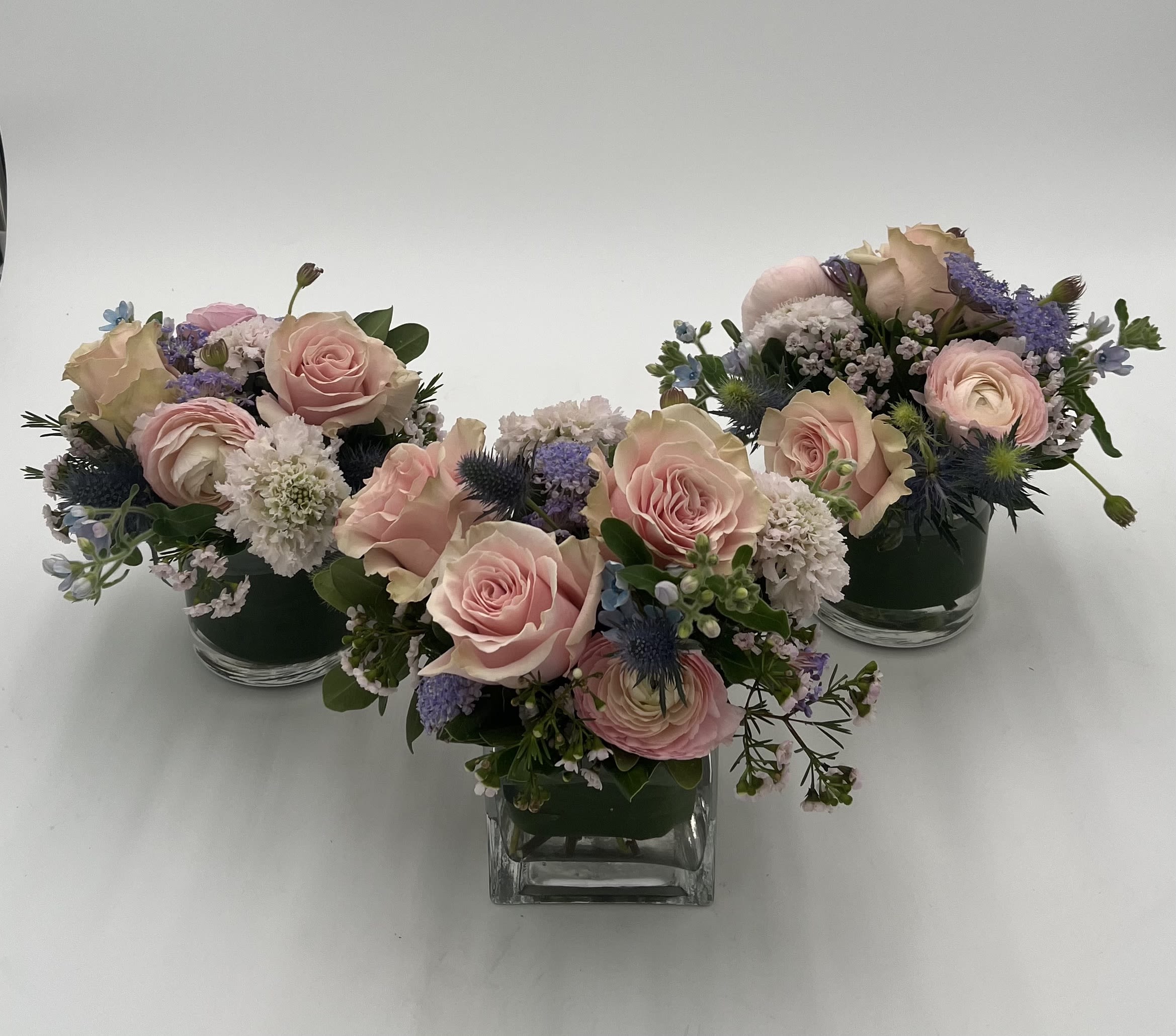 Petit Trio - (3) Petite arrangements that may be grouped together or placed in different locations.  Also, looks great as a dining table centerpiece.  Rose, thistle,  ranunculus, wax flower, ranunculus and scabiosa in shades of pink and lavender. 