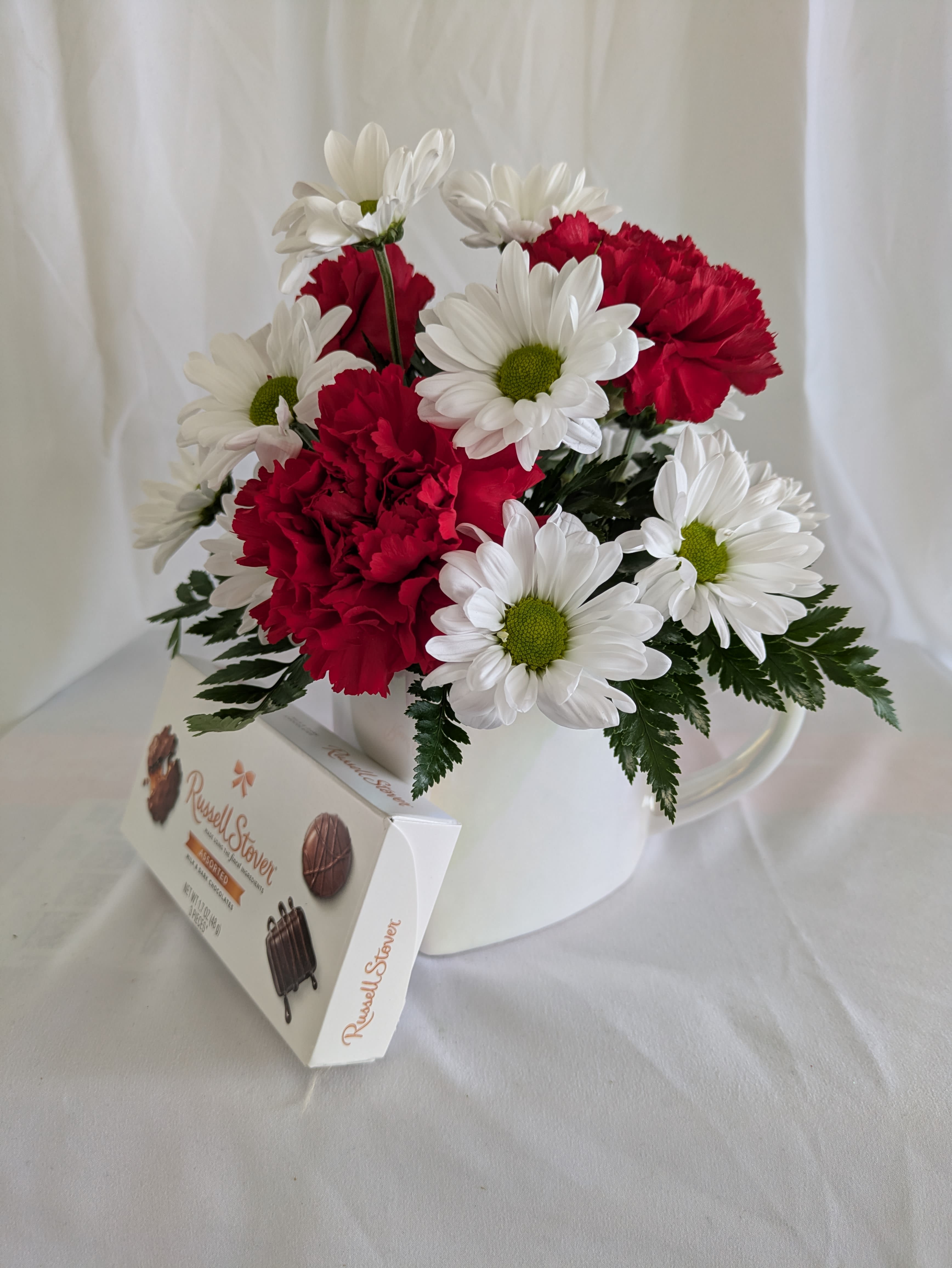 SWEET SENTIMENTS - A CHARMING MIX OF RICH RED CARNATIONS AN CHEERFUL WHITE DAISIES, THOUGHTFULLY ARRANGED IN A KEEPSAKE MUG AND PAIRED WITH A SWEET CHOCOLATE TREAT. THIS COZY DESIGN IS PERFECT FOR SHARING LOVE, COMFORT, AND A LITTLE INDULGENC ALL IN ONE THOUGHFUL GIFT.