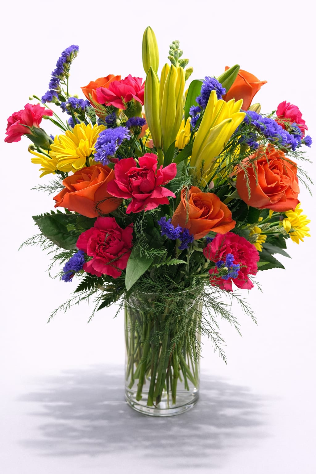 Vibrant Celebration - A bold and joyful floral arrangement designed in a clear glass cylinder vase, featuring a lively mix of orange roses, hot pink carnations, yellow lilies, yellow daisies, and purple statice, accented with fresh greenery.  This design is full of energy and movement, combining warm and vibrant tones that symbolize happiness, celebration, and positivity. The unopened lilies add height and elegance, allowing the arrangement to continue blooming beautifully over time.  Perfect for birthdays, congratulations, anniversaries, get-well wishes, celebrations, and cheerful home décor, this arrangement is made to brighten any space instantly.