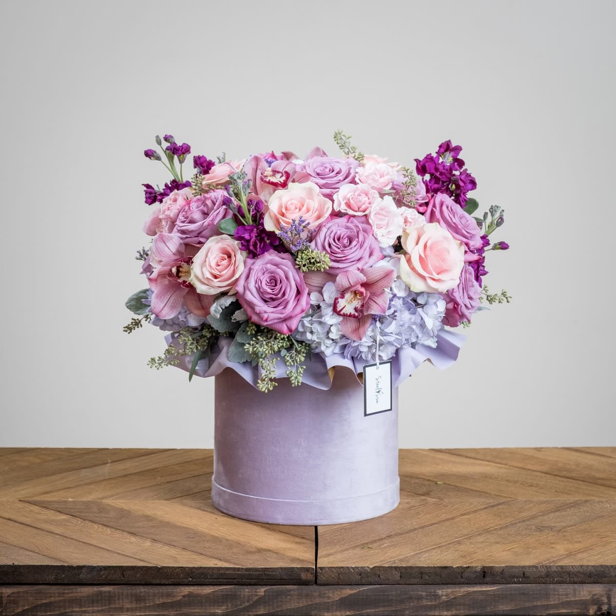 Babe - A dusty lavender velvet cylinder box is flourishing with out favorite pastels. A mixture of light pink and lavender roses, lavender hydrangea blooms, playfully purple stems of stock, tropical cymbidium orchid blooms, purple spray roses, and complimented with dusty miller and seeded eucalyptus.  Measures approx. 16" x 16"