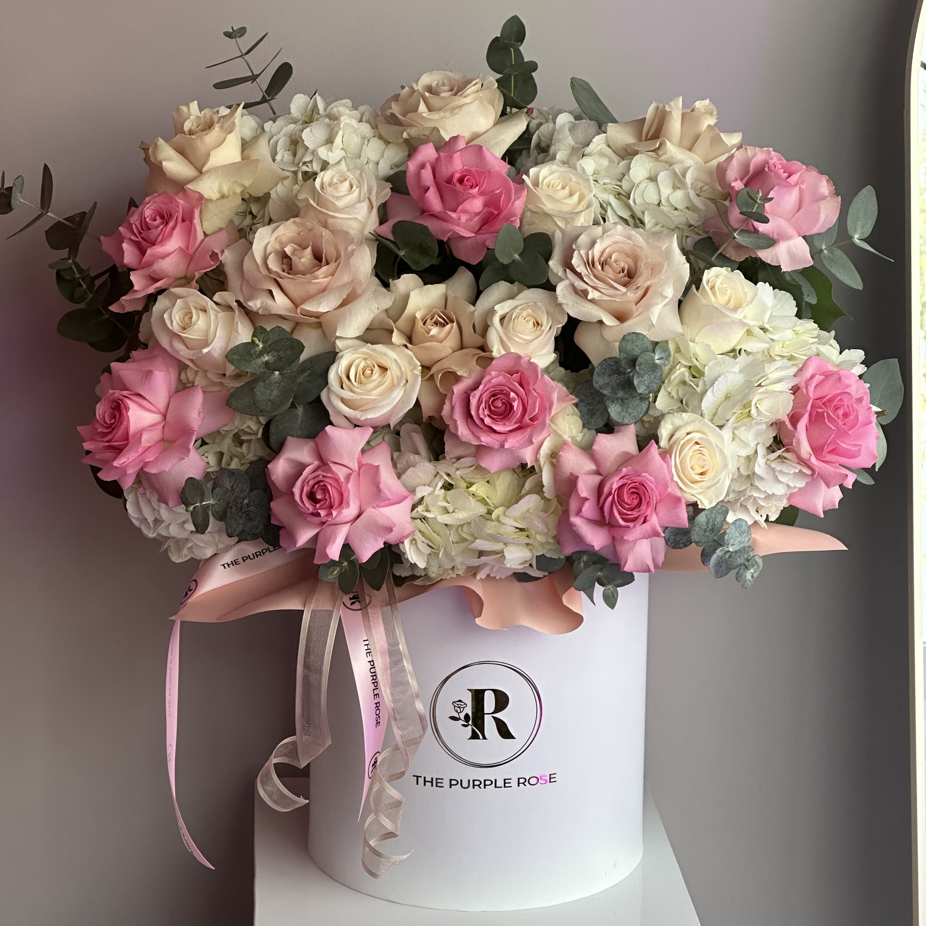 Pink Champagne - A full and elegant arrangement featuring pink and ivory roses, white hydrangeas, and soft greenery designed in our signature hatbox.  DELUXE size is in the main picture.