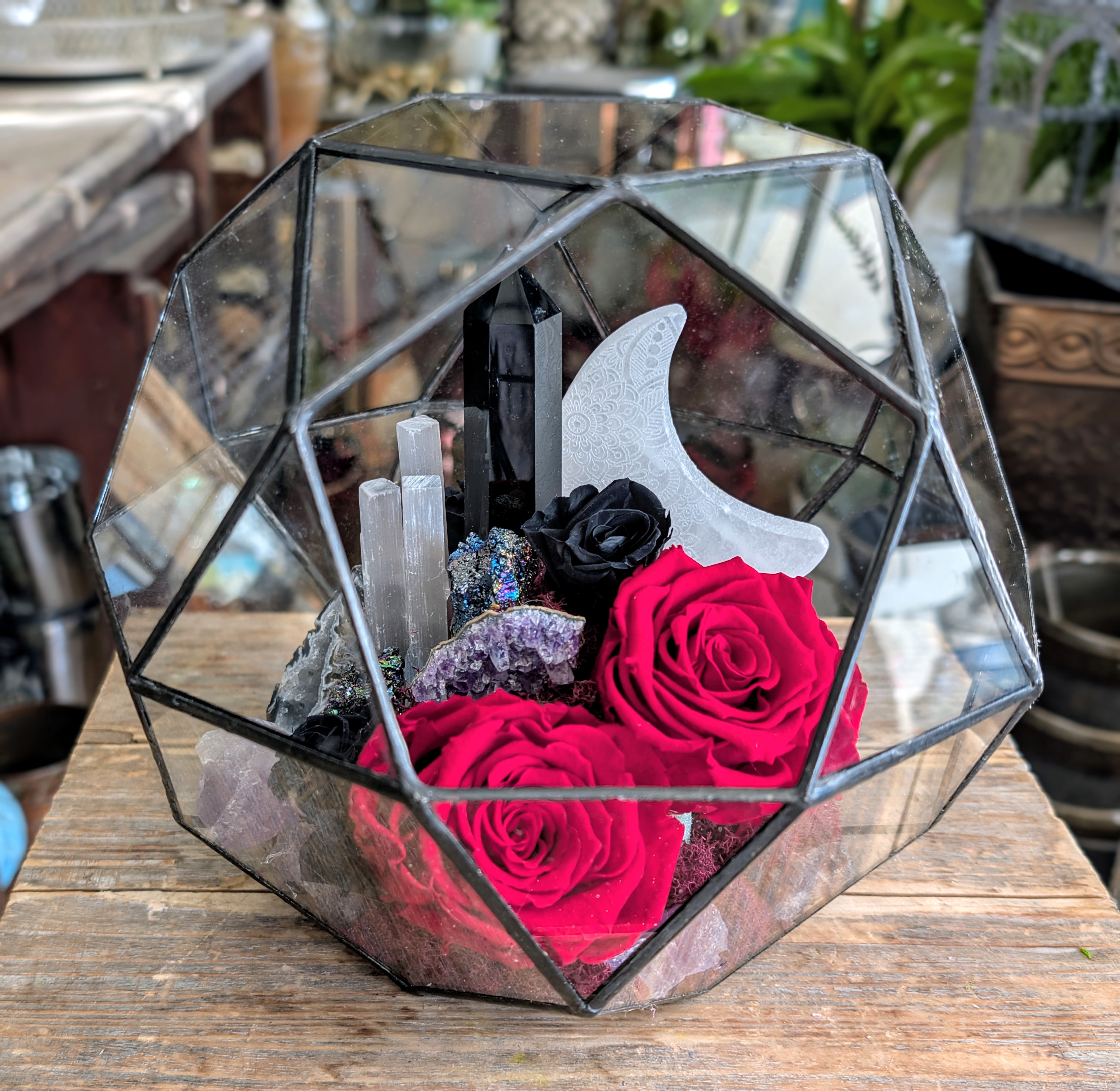 Obsidian -A Preserved Rose And Crystal Arrangement  - Dark, powerful, and incredibly romantic, Obsidian is a striking preserved floral and crystal arrangement designed to feel like a modern relic—equal parts luxury and mysticism. Velvety preserved red roses anchor the design, their rich crimson tones symbolizing passion and devotion, while a miniature black roses add depth, contrast, and a touch of the arcane. Nestled within a faceted geometric glass terrarium, shimmering crystal elements—including obsidian-inspired accents, amethyst textures, and luminous selenite forms—create a sculptural landscape that feels celestial and grounded all at once. A crescent moon detail rises softly among the florals, lending an ethereal, lunar energy to this one-of-a-kind piece. Perfect for Valentine’s Day, anniversaries, or meaningful moments that call for something bold and unforgettable, Obsidian is crafted using preserved botanicals for long-lasting beauty with no maintenance required. This arrangement is designed to be both a floral statement and an art object—timeless, dramatic, and deeply intentional. Created by Windmill Floral Expressions, this luxury preserved arrangement is ideal for clients seeking a distinctive Las Vegas florist offering custom, artistic designs with a modern, moody edge. Available for delivery in Las Vegas, Henderson, and surrounding areas. A love story written in shadow and light.