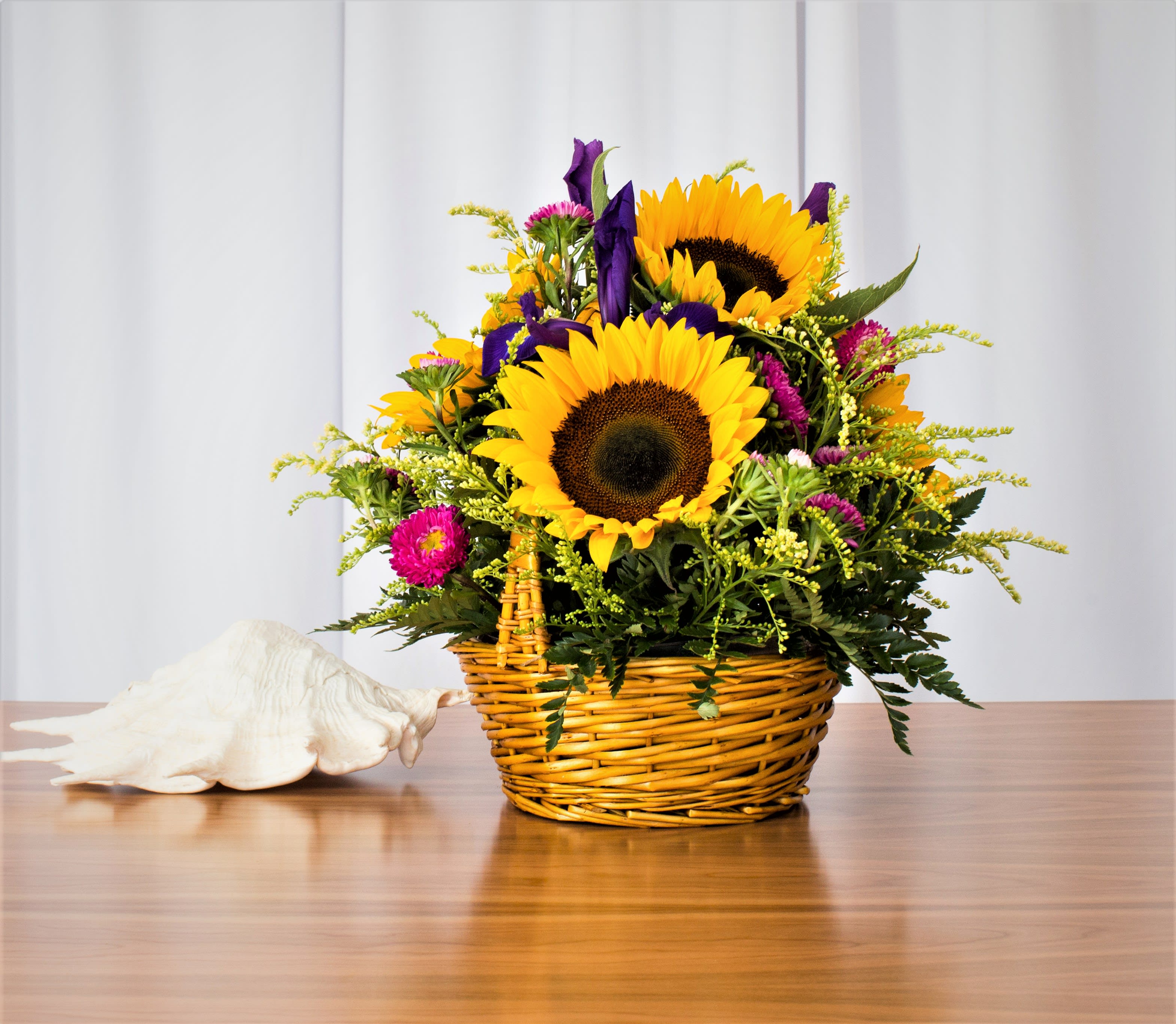 Happy Wishes Basket  - Brighten anyone's day with this delightful basket featuring sunflowers, asters, and more. Perfect for any setting, including dining tables, coffee tables, or desks, this cheerful arrangement brings warmth and joy to every space.  Key Features:  Cheerful Sunflowers and Asters: Showcases vibrant sunflowers and charming asters, complemented by a variety of seasonal blooms.  Versatile Display: Ideal for enhancing dining tables, coffee tables, desks, or any area that needs a touch of brightness and cheer.  Beautiful Basket: Arranged in a lovely basket that adds rustic charm and makes the bouquet easy to display anywhere.  Perfect for Any Occasion: Whether as a gift or a decor piece, this arrangement is sure to bring smiles and brighten up any setting.  Add a touch of sunshine to your space with our Bright Sunflowers and Asters Basket, and enjoy the uplifting beauty it brings to every corner of your home or office.       