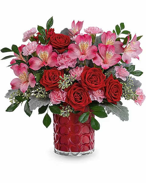 ENCHANTED HEARTS BOUQUET - Adorned with a charming heart pattern and gleaming with a pearlescent finish, this delightful glass vase adds an extra touch of magic to their Valentine's Day when filled with a stunning red and pink bouquet. This Valentine's Day arrangement features red spray roses, pink alstroemeria, and miniature light pink carnations, complemented by dusty miller, seeded eucalyptus, and huckleberry. This vibrant red and pink bouquet is delivered in the Enchanted Hearts vase.  