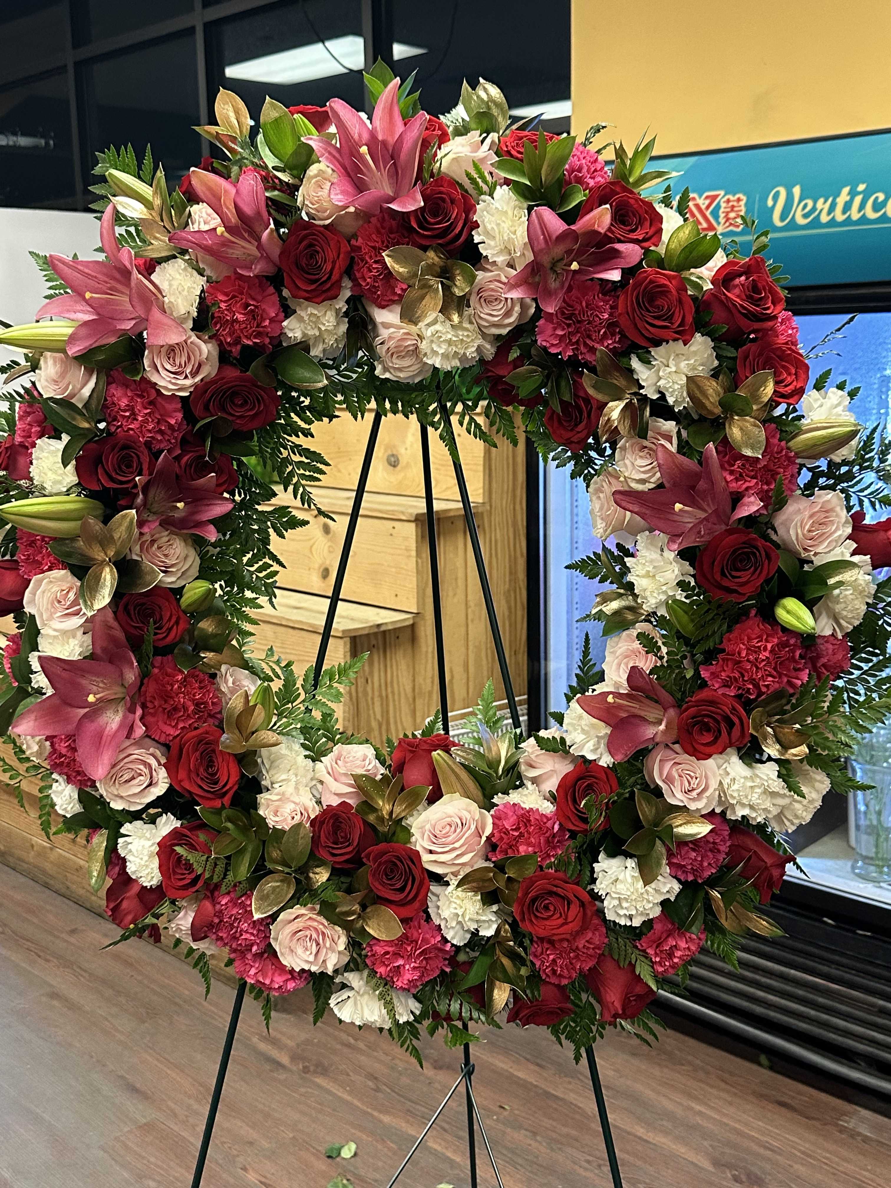 PINK, RED, AND WHITE (DESIGNER CHOICE WREATH) - Standard Size is an 18 inch wreath with 25 roses and other flowers Deluxe Size is a 22 inch wreath with 36 Roses and other flowers Premium size is a 24inch wreath with 42 roses and other flowers  WREATH INCLUDES A MIXTURE OF PINK , RED AND WHITE FLOWERS.  THIS WREATH HAS A MIXTURE OF ROSES , CARNATIONS, AND LILIES AND GOLD RUSCUS LEAVES.  THIS ARRANGEMENT INCLUDES THE ROSES STATED PER PRICE.