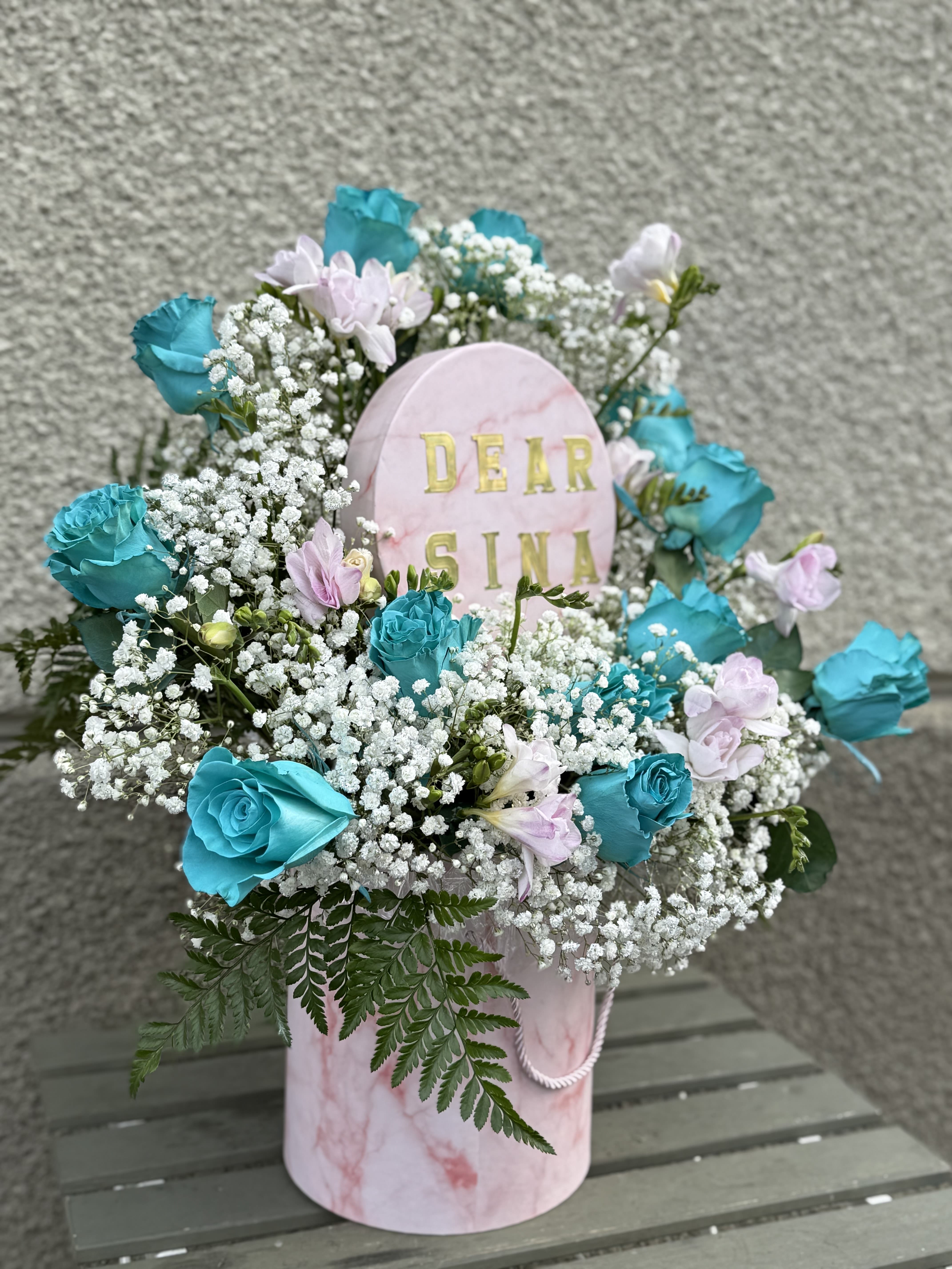 ROUND BOX - **Floral Arrangement Description**  This elegant floral arrangement showcases vibrant turquoise roses paired with soft blush pink blooms, delicately accented by clouds of white baby’s breath. Lush green foliage adds natural texture and balance, creating a fresh and graceful presentation. Designed in a blush marble-style hat box with a subtle rope handle, this arrangement blends modern charm with timeless beauty—perfect for birthdays, anniversaries, congratulations, or simply to brighten someone’s day. 