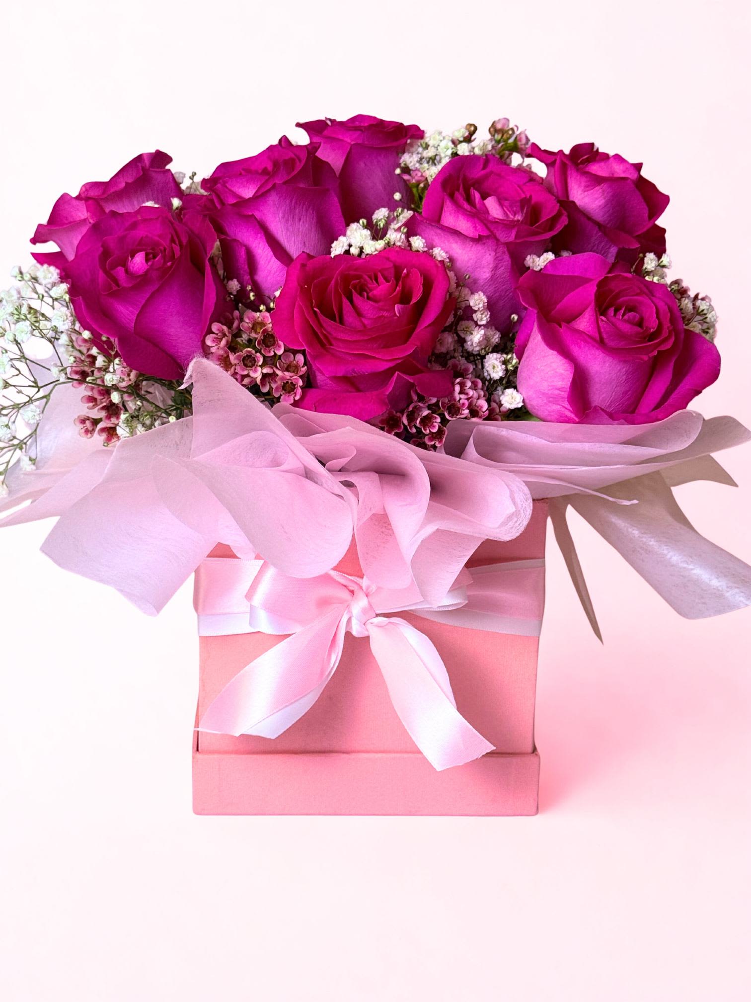 Enchanted Pink Rose Box - At the center is a lush bouquet of one dozen pink roses, each bloom full and velvety, symbolizing love and passion. The roses are tightly arranged to create a rich, rounded shape, with fresh green foliage peeking through for contrast. They’re wrapped in elegant paper and placed in a sleek box, finished with a satin red ribbon for that timeless Valentine’s touch.  The whole arrangement gives off a “wow” moment—romantic, thoughtful, and heartfelt—perfect for saying I love you in a way that feels both meaningful and memorable. 