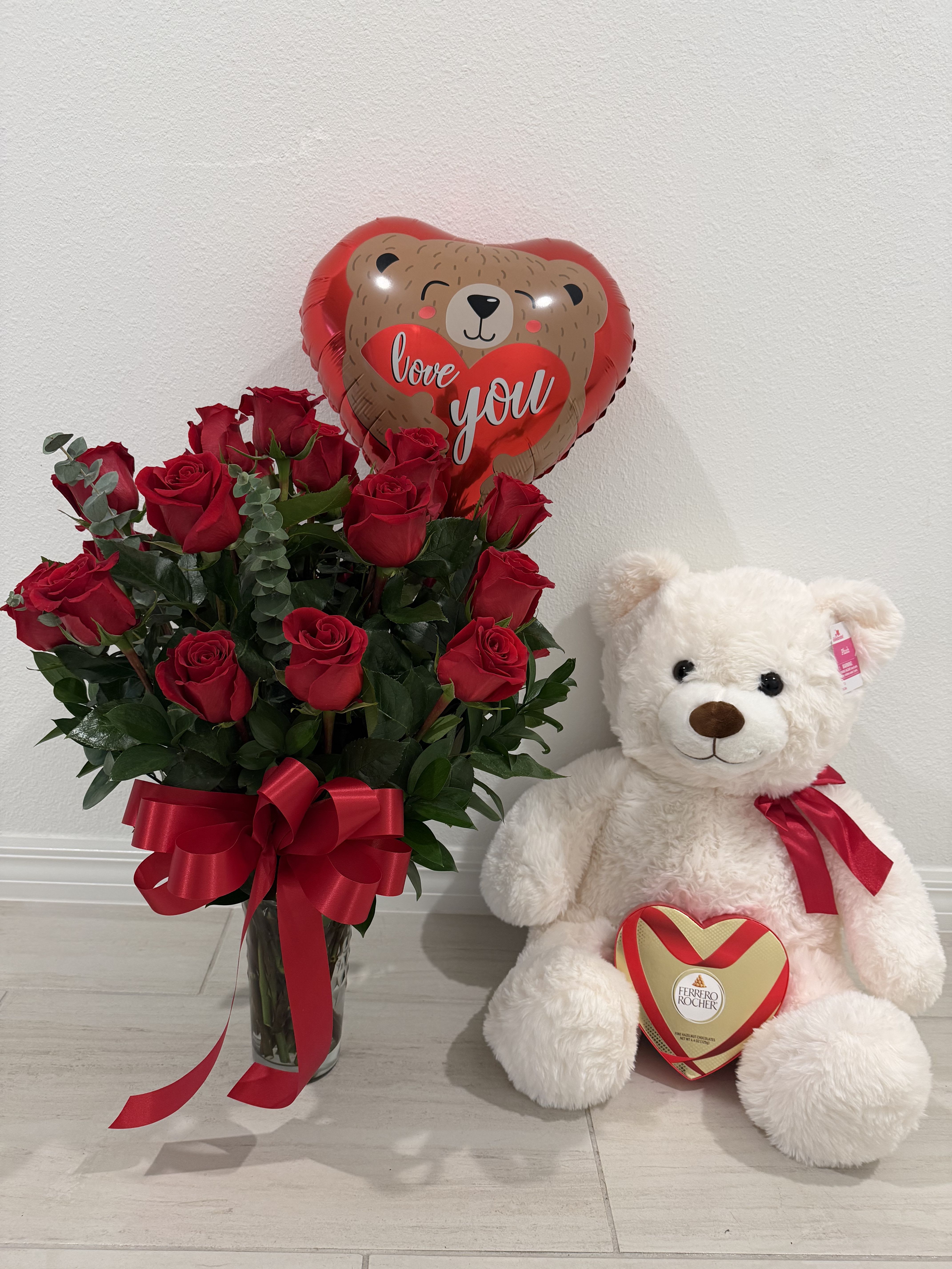 Forever Love Roses &amp; Teddy Gift Set - A classic romantic gift set featuring a stunning arrangement of fresh red roses in a clear glass vase, accented with lush greenery and finished with a red satin bow. Topped with a heart-shaped “Love You” balloon, this arrangement is paired with an adorable plush teddy bear holding a Ferrero Rocher heart-shaped chocolate box.