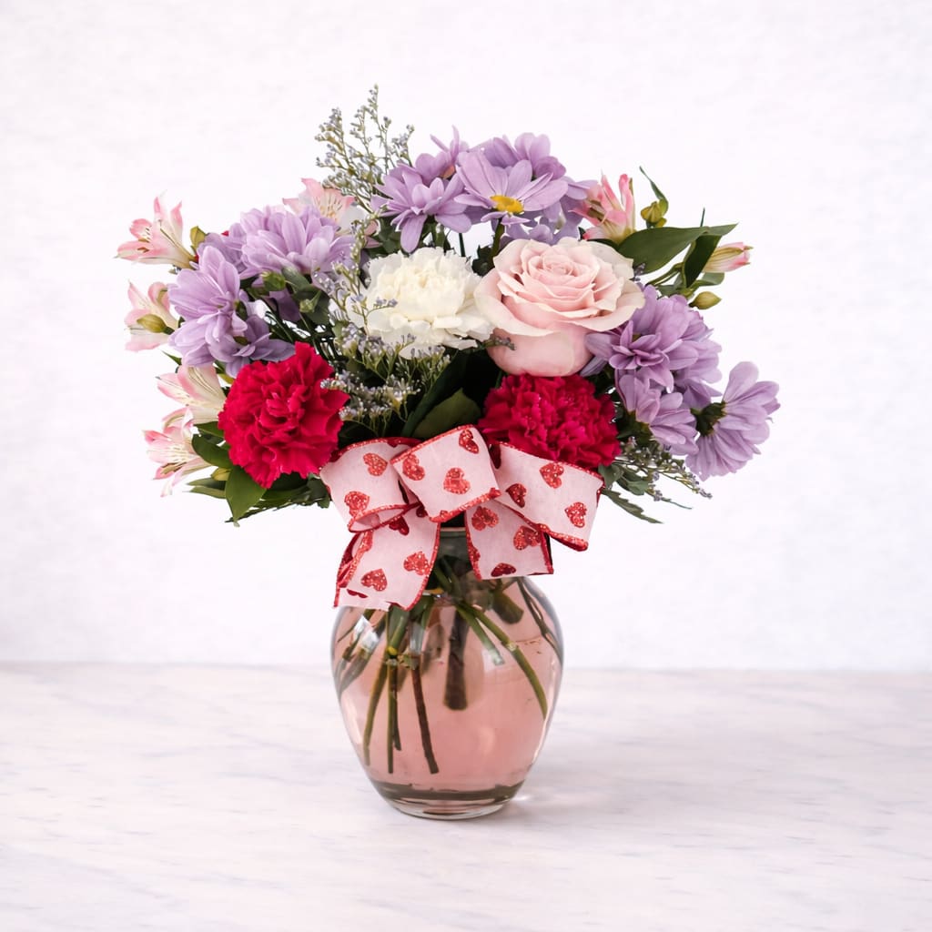 REMIX - REMIX is a soft and cheerful arrangement featuring vibrant red carnations, blush pink roses, creamy white blooms, and delicate lavender accents. Designed in a rosy pink glass vase and finished with a heart-patterned ribbon, this arrangement is perfect for Valentine’s Day, anniversaries, or sending a sweet reminder of love and appreciation.
