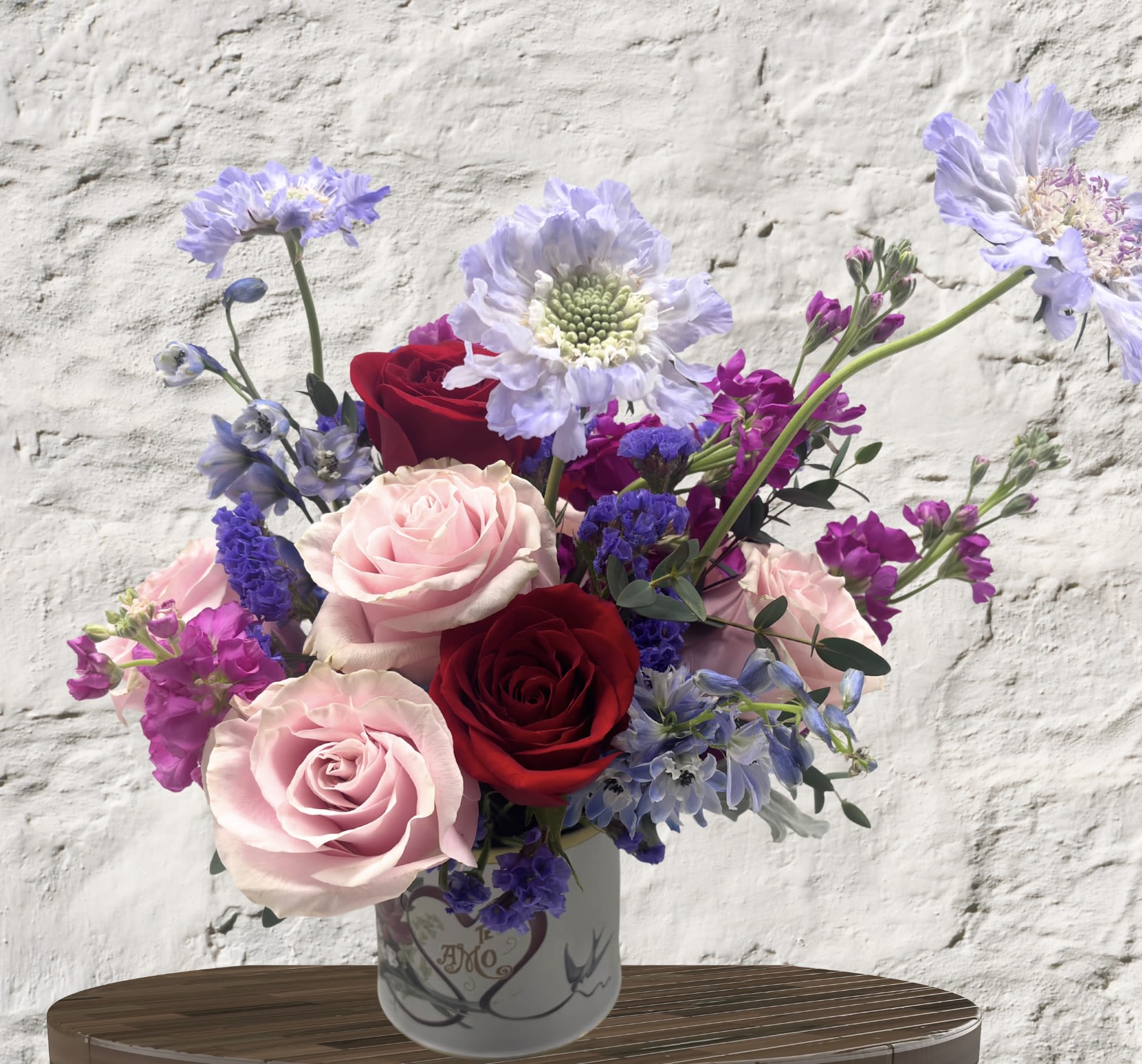 Amor en Flor - Celebrate love in every language with this striking, soulful arrangement. Deep crimson roses symbolize passion, while soft pink blooms and delicate lavender scabiosa add a touch of gentle romance.  This multi-dimensional bouquet is layered with vibrant purple statice, wispy delphinium, and lush magenta stocks, creating a rich tapestry of color. Nestled in a charming "Te Amo" metal vessel, it is a heartfelt statement piece perfect for anniversaries, Valentine's Day, or simply saying "I love you" with a flourish of garden elegance