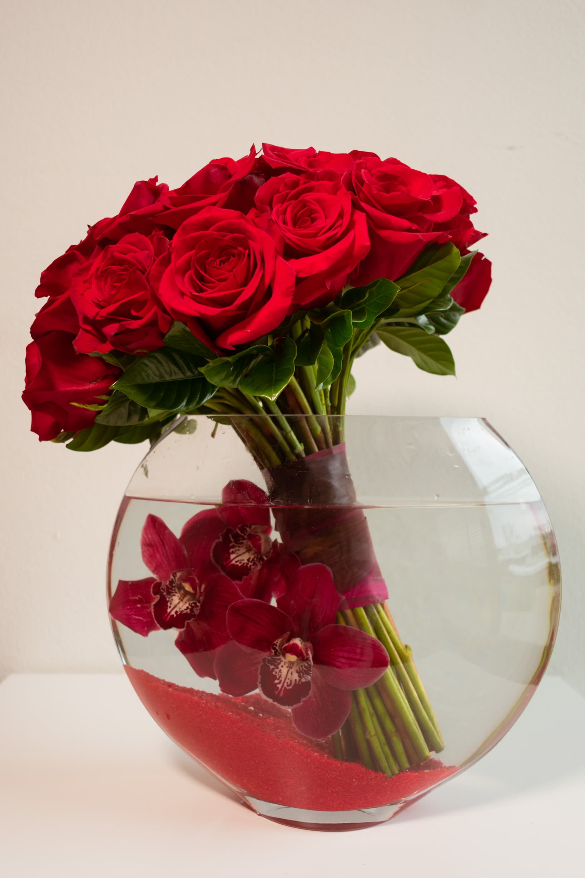 Endless Romance - Love reimagined through the art of floral design...  Prepare to be mesmerized by this show-stopping arrangement that transforms the classic red rose bouquet into a work of contemporary art. Two dozen premium red roses crown a crystal-clear fishbowl vase, their stems elegantly bundled and submerged alongside burgundy cymbidium orchids that seem to dance beneath the surface of the water like captured poetry. This is romance with a modern twist—sophisticated, unexpected, and absolutely unforgettable.  Perfect For: -Anniversary celebrations that deserve something extraordinary - Romantic proposals seeking the element of surprise   - Valentine's Day gifts that go beyond the expected - Modern home decor with contemporary flair - Corporate gifts that make a lasting impression - Date night centerpieces that spark conversation   -Height: Approximately 16 inches total 