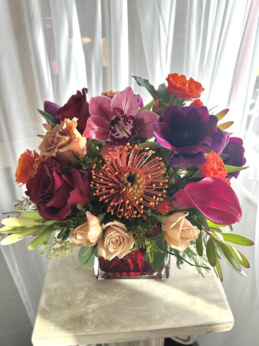 La Isla Bonita - Purple anemoneas, red or hot pink roses, pin-cushion proteas and more are artfully arranged in a red cube vase.  Call the shop to find out exactly what we will be using because our inventory changes daily.