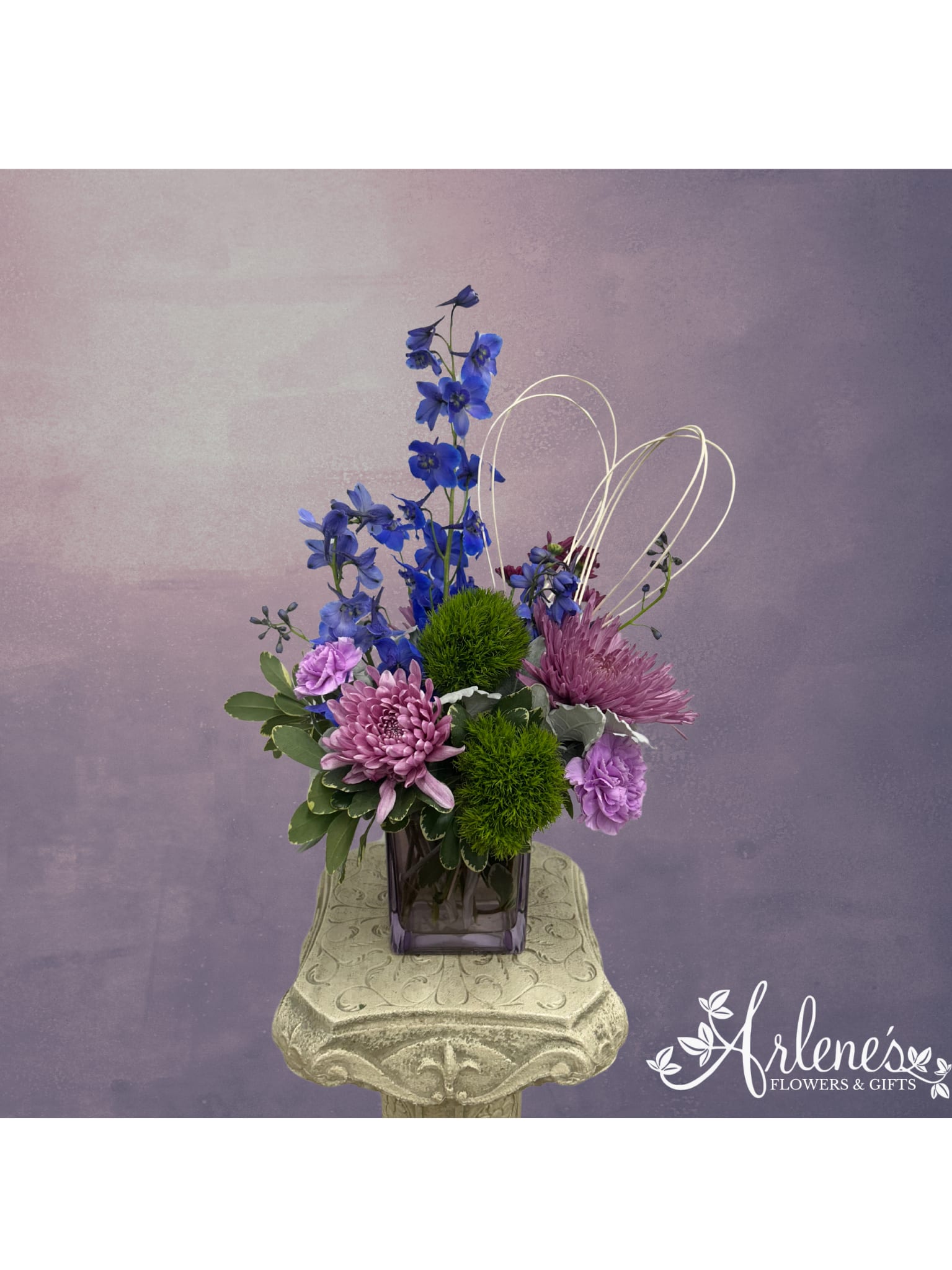 Country Charm - This purple valentine country floral arrangement combines rustic charm of the countryside with the romantic lures of Valentine's day! This arrangement features blue delphinium, purple cremons, green dianthus balls and purple daises. 