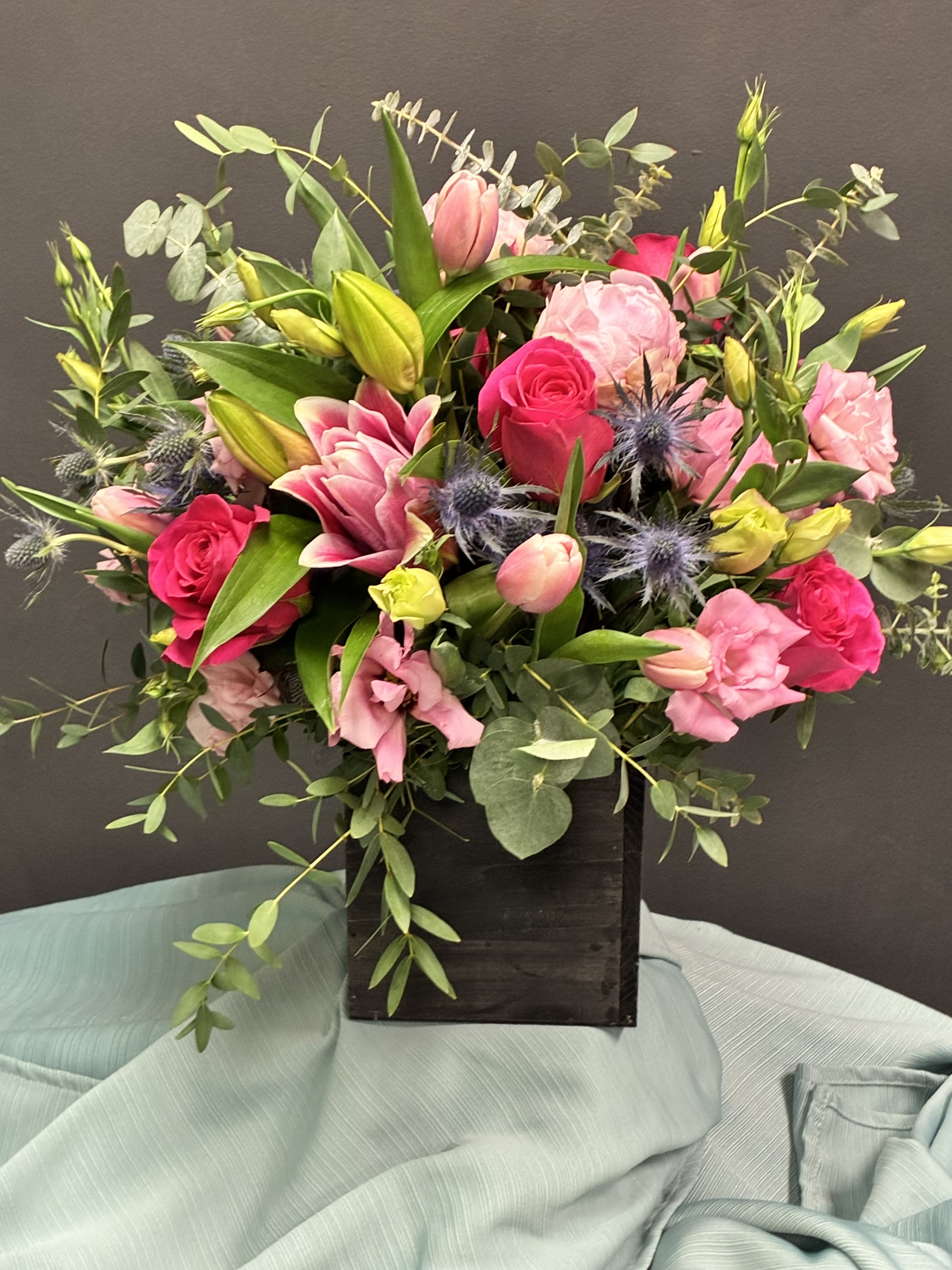 Pink Reverie - A lush celebration of pink blooms and bold textures, this arrangement features a graceful medley of roses, lilies, tulips, and carnations in soft blush and vibrant fuchsia tones. Spiky blue thistles add a striking contrast, while green buds and eucalyptus leaves lend freshness and movement. The flowers rise from a sleek black rectangular vase, grounded on a draped light blue fabric that enhances the elegance of the display. This bouquet radiates romance, sophistication, and a touch of wild charm—perfect for celebratory moments, heartfelt gestures, or modern décor.