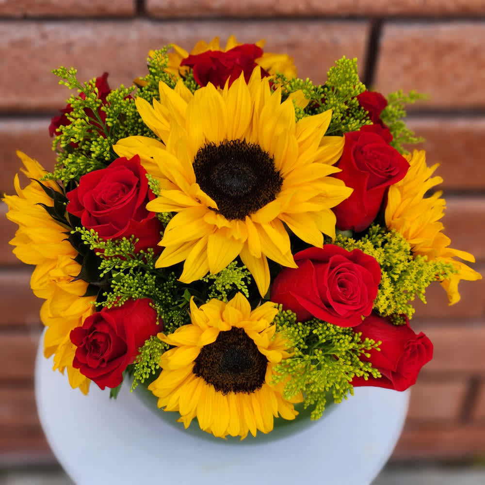 Red Roses and Sunflowers Bowl - It's romantic. It's sunny. It's gorgeous! Ecuadorian Red Roses and Sunflowers are an ideal mix for sending both love and cheer. This bouquet is presented in a glass bubble bowl vase lined with a tropical leaf and colored beads. Standard size is approximately 10in (W) x 10in (H). Deluxe version is larger and features more blooms hand arranged in a larger bowl vase.  Standard - 8 Red Roses, 5 Sunflowers, Seasonal Filler and Fresh Garden Greens - 8in Bubble Bowl Vase  Deluxe - 12 Red Roses, 10 Sunflowers, Seasonal Filler and Fresh Garden Greens - 10in Bubble Bowl Vase  Care Tips: Place your bouquet in a cool location. Don't put the arrangement in direct sunlight, near heating or cooling vents, in drafty places, directly under ceiling fans, or on top of televisions or radiators. Check water level daily, keep the vase full with clean water. Change water every 2-3 days and apply a sharp fresh cut to the stems. This process will ensure extended flower's life span.