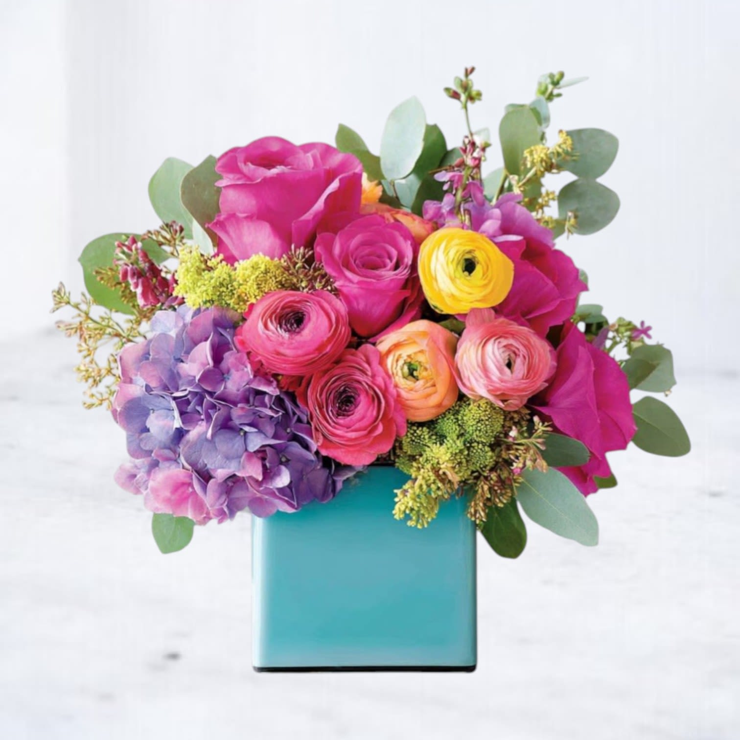 Vivid Bloom - Vibrant, bold, and full of life — Vivid Bloom is a celebration in floral form. Bursting with vivid fuchsia roses, sunny yellow and orange ranunculus, rich purple hydrangea, and lush greenery, this energetic arrangement makes a confident statement in its modern turquoise cube vase. Whether you're celebrating a birthday, sending congratulations, or brightening someone’s day “just because,” this bouquet is sure to deliver joy.  Substitution Policy: In the event that certain blooms or containers are unavailable, Twigs and Blooms will make thoughtful substitutions with flowers and vessels of similar color, style, and quality. Cymbidium orchids, while striking, may occasionally be replaced with another premium focal bloom like garden roses or calla lilies. Vase substitutions will always maintain a clean, modern look unless otherwise requested.