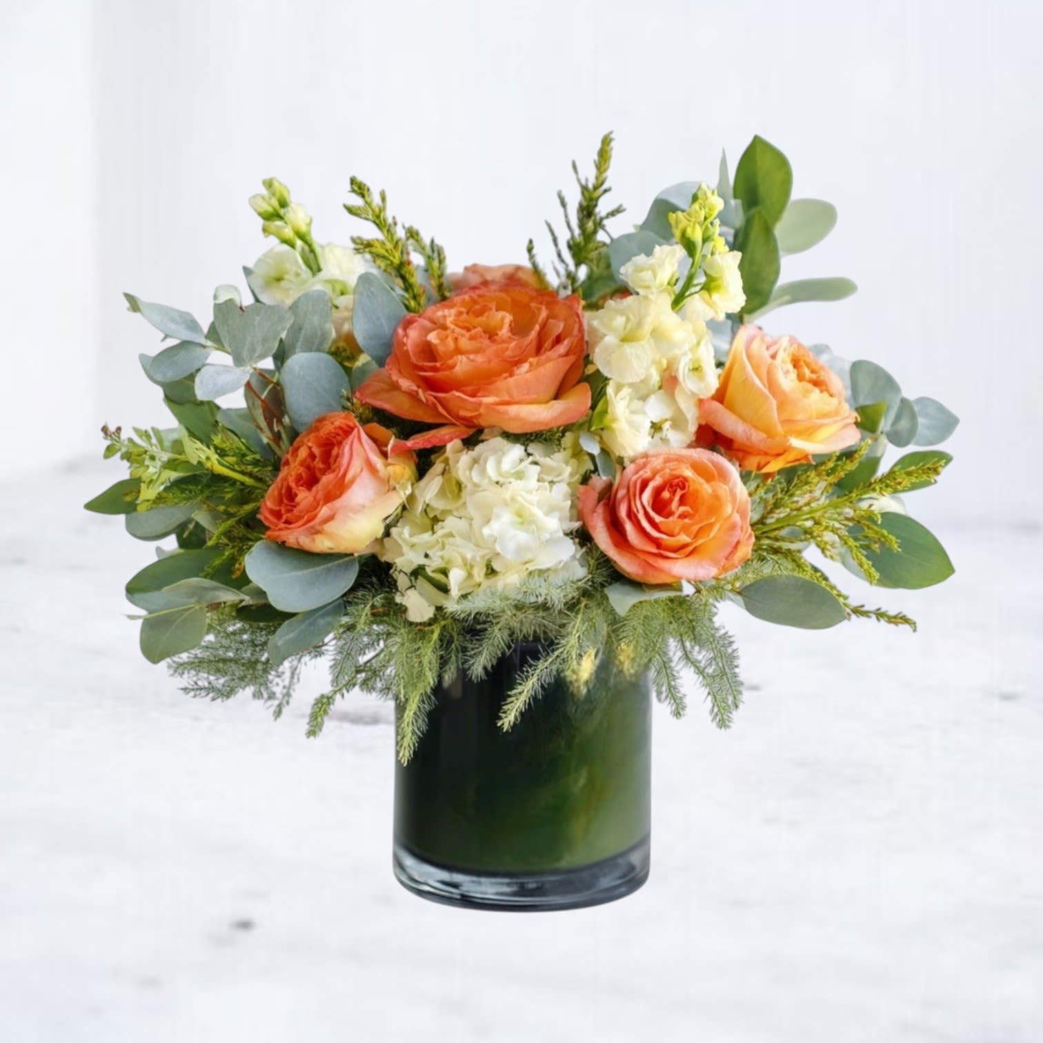 Citrus Grove Luxe - Capture the essence of a sun-drenched citrus grove with this lush and textural arrangement. Citrus Grove Luxe features a radiant blend of orange, peach, and coral roses paired with creamy white blooms and cascading eucalyptus. Designed in a modern glass cylinder vase lined with fresh foliage, this bouquet brings effortless elegance and warmth to any space.  Perfect for everyday occasions, birthday celebrations, thank-you gifts, or as a sophisticated statement piece for homes and offices in Galveston, South Houston, and nearby coastal communities. Local delivery available.  Substitution Policy: Our arrangements are crafted fresh daily using the season’s best blooms. In the rare event that specific flowers (such as stock, peach roses, or filler accents) are unavailable, we will thoughtfully substitute with blooms of equal value and similar color tone to maintain the design and style. Container may vary slightly based on availability.