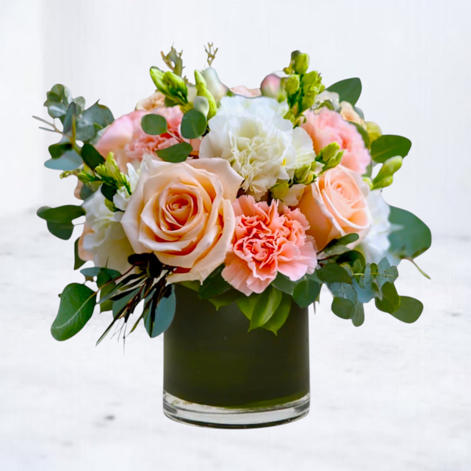 Eleanor - A soft, romantic floral arrangement designed with natural movement and gentle elegance. Featuring peach roses, blush carnations, creamy white blooms, and fresh eucalyptus, this design is airy, balanced, and thoughtfully composed. Arranged in a clear glass vase, it offers a timeless garden-inspired look with warm, inviting tones.  Perfect for birthdays, anniversaries, thank-you gifts, sympathy gestures, or simply sending something beautiful just because.  Flower varieties may vary slightly based on seasonal availability while preserving the overall color palette and design style.