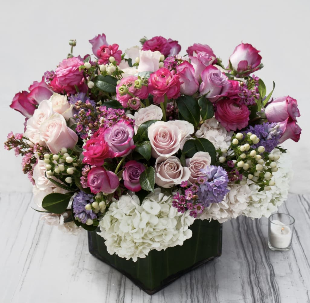 Powerful Pastels (DB) - Prepare to be enchanted by our magnificent floral arrangement, a stunning display of pink and lavender roses, white hydrangea, pink ranunculus, white hypericum, and more. This opulent creation is expertly arranged in a large leaf-lined glass cube, creating a masterpiece that exudes elegance and beauty.