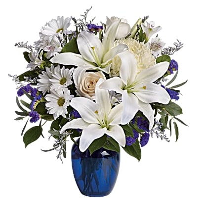 Blue Skies - Blooms such as crème roses, graceful white oriental lilies, white alstroemeria, a white disbud mum, purple statice and lavender limonium are accented by seeded eucalyptus and salal in a stunning cobalt blue glass vase..