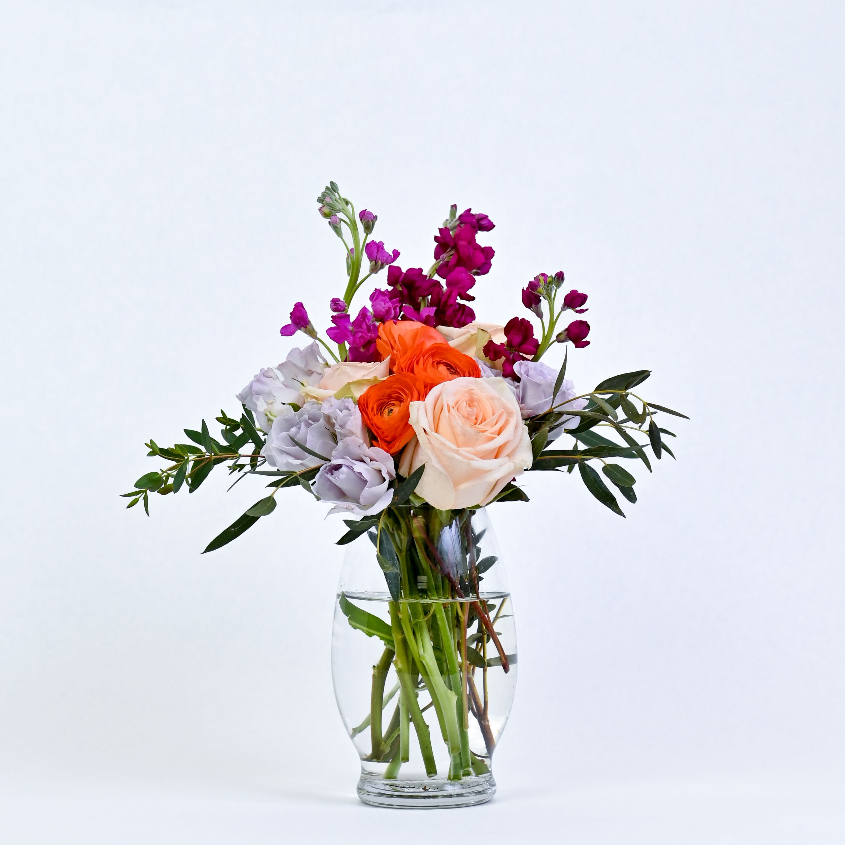 Sweet Garden Glow - A vibrant, garden-inspired arrangement featuring lavendar lisianthus, orange ranunculus, fuchsia stock, soft pink roses, and fresh gunni- beautifully blended for a cheerful, colorful display full of charm. 