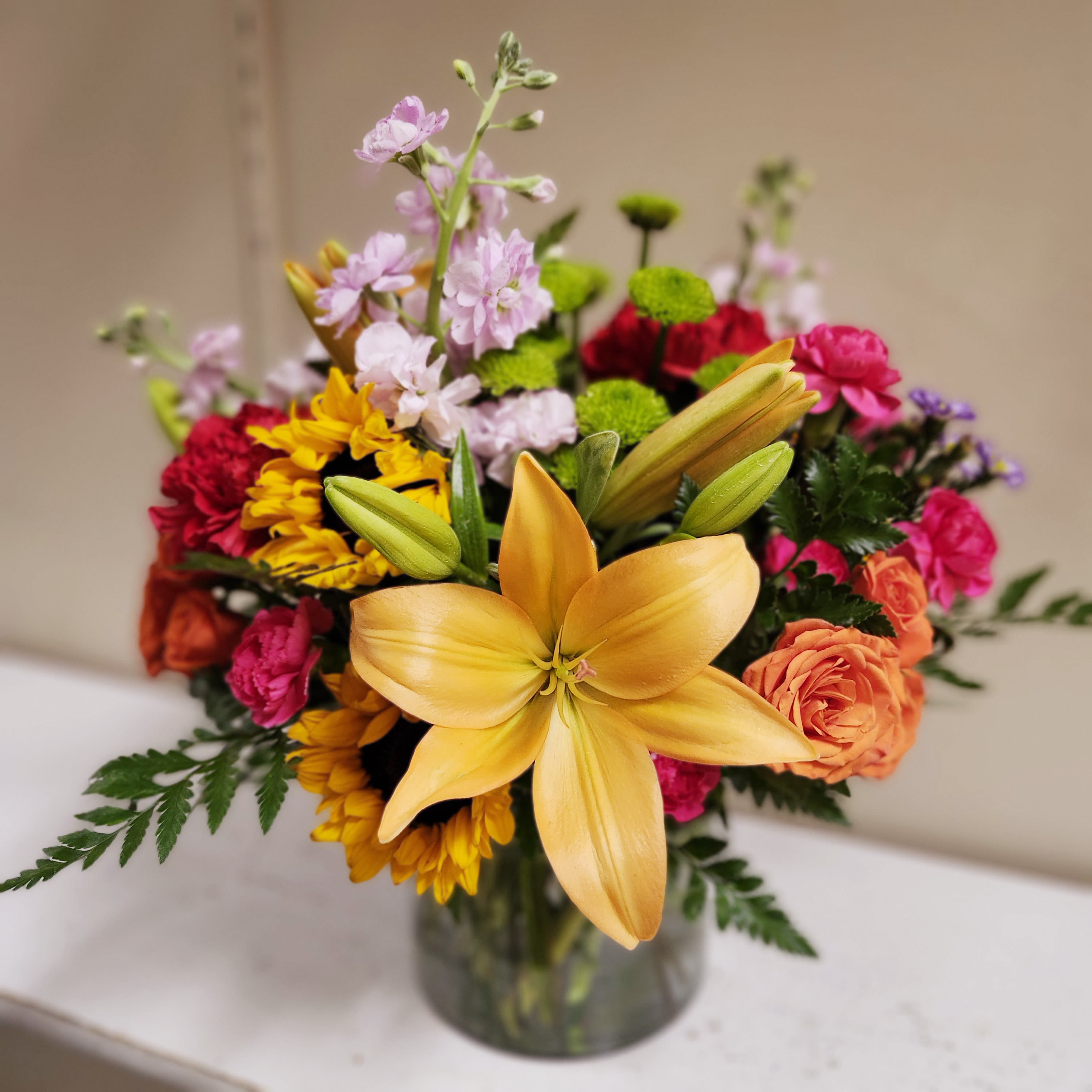 Sunshine Symphony by Latin Lady Flowers - Brighten any room with the Sunshine Symphony floral arrangement, a vibrant ensemble of large yellow lilies, orange spray roses, pink mini carnations, purple stock flowers, green button poms, and yellow sunflowers. Expertly arranged in a clear glass vase, this lush and colorful bouquet brings warmth and joy to any occasion. Perfect for birthdays, anniversaries, or simply to make someone smile. 