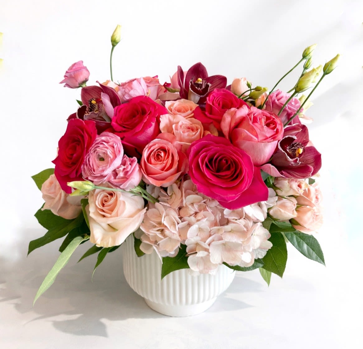 Heartbreaker - A lush celebration of love in full bloom with vibrant hot pinks, soft blush tones, and cream whites for a look that’s playful, romantic, and irresistibly eye-catching. Featured flowers include premium garden roses, Italian ranunculus, cymbidium orchids, lisianthus, and hydrangeas in a beautiful, ribbed ceramic vase.