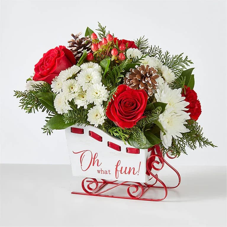 Sleigh Bells Bouquet - Sled into the holiday season with this lush arrangement of red roses, carnations and fragrant greens. This keepsake sleigh is perfect for a centerpiece, party décor, hostess gifts, or to send holiday wishes to those far away!