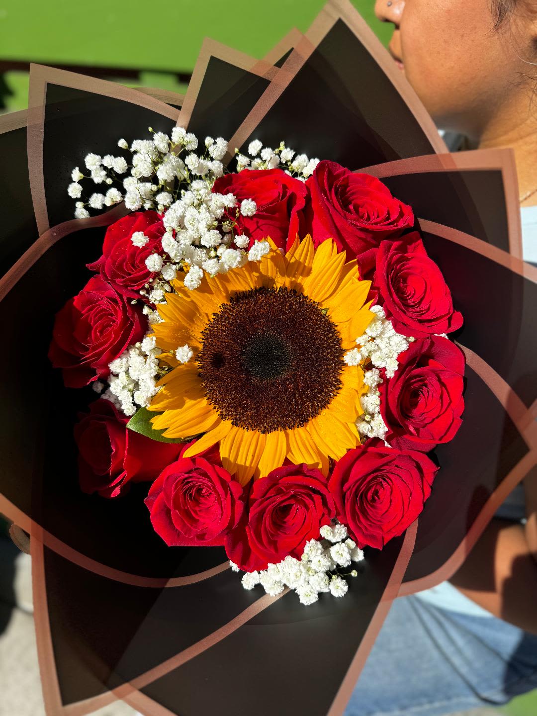 Golden Sunburst Bouquet - A striking bouquet featuring a radiant sunflower at the center, surrounded by lush red roses and delicate baby's breath, all wrapped in an elegant dark-toned design. This vibrant arrangement symbolizes warmth, love, and happiness—perfect for brightening someone's day
