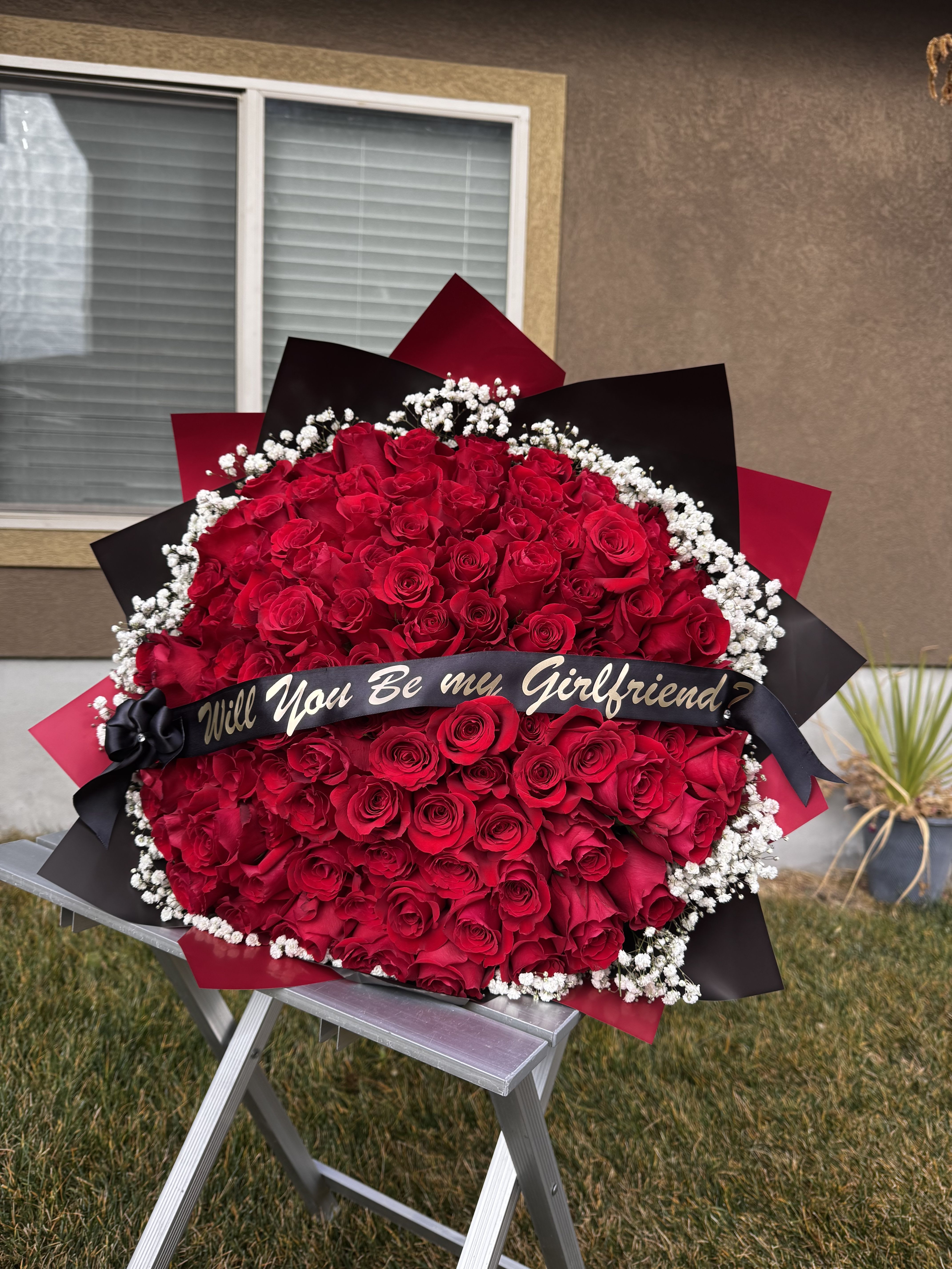 “Will You?” – Signature 100 Roses Bouquet - A bold, unforgettable bouquet designed for life-changing moments.  This luxury arrangement features 100 premium red roses, framed with delicate gypsophila, elegant wrapping, and a custom banner that asks the question that matters most: “Will you be my girlfriend? Or Will you be my Valentine?  Perfect for Valentine’s Day, proposals, or romantic gestures that deserve to be remembered forever. This is not just flowers — it’s a statement.  ️ 100 red roses ️ Gypsophila (baby’s breath) ️ Luxury wrapping (black &amp; red) ️ Custom banner included ️ Handcrafted with intention and detail
