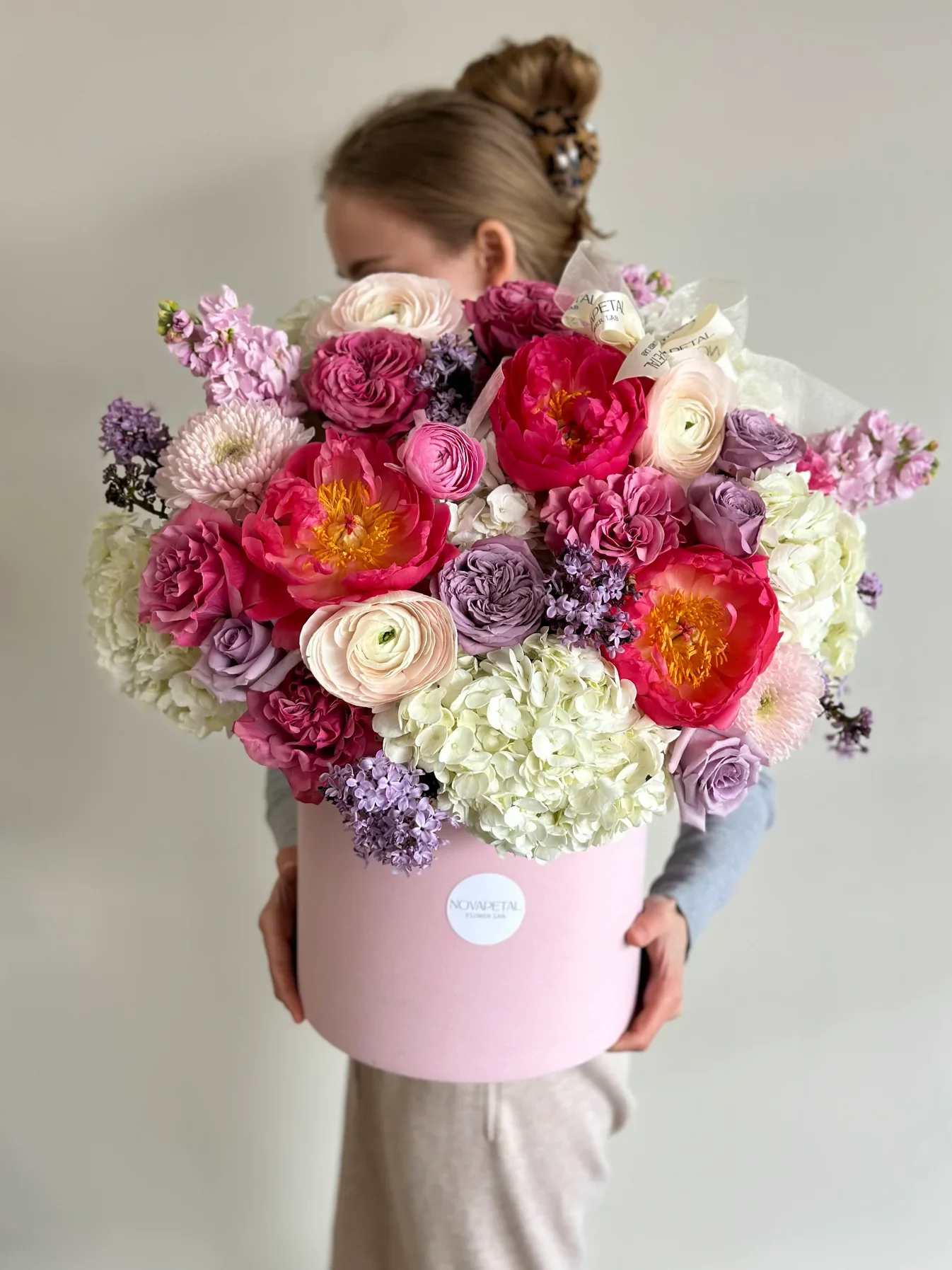 Lilu box arrangement - This exquisite floral arrangement is a stunning composition that exudes luxury and elegance. Presented in a soft pink, round box that adds a touch of modern sophistication, the bouquet is a rich tapestry of colors and textures. It features an abundant collection of flowers including lush peonies in shades of bright pink, cream, and soft lavender roses, interspersed with delicate hydrangea blooms that provide a dense, cloud-like foundation. The arrangement is thoughtfully accented with ruffled ranunculus and hints of lilac-hued flowers, contributing to the overall pastel palette that makes this bouquet a visual delight. The flowers are carefully selected for their premium quality and arranged to create a harmonious balance, perfect for making a statement on any occasion.  Please note that due to seasonal availability of flowers, some blooms in the arrangements may be substituted for a flower of equal beauty and quality. Our expert designers ensure that the overall aesthetic and color scheme of the bouquet is maintained.  Pictured: Large box  Last picture is a Medium