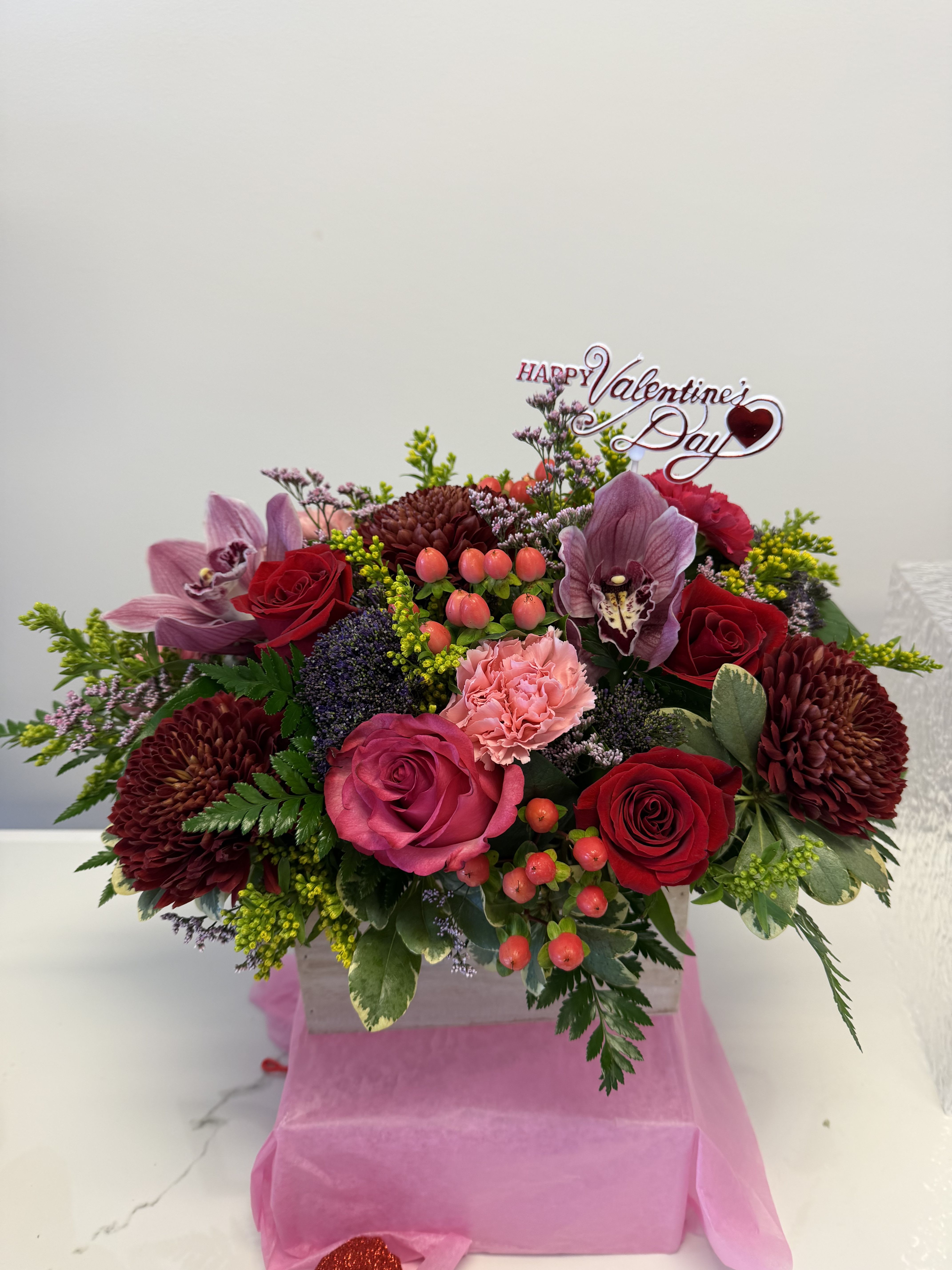 Valentine’s centrepiece - Celebrate love in full bloom with this luxurious Valentine’s Day floral arrangement. Designed in a charming wooden box, this elegant piece features rich red and pink roses, exotic cymbidium orchids, lush burgundy mums, soft pink carnations, and seasonal accents of hypericum berries and textured greenery. Finished with a delicate “Happy Valentine’s Day” topper, it’s a statement of romance, warmth, and timeless beauty.