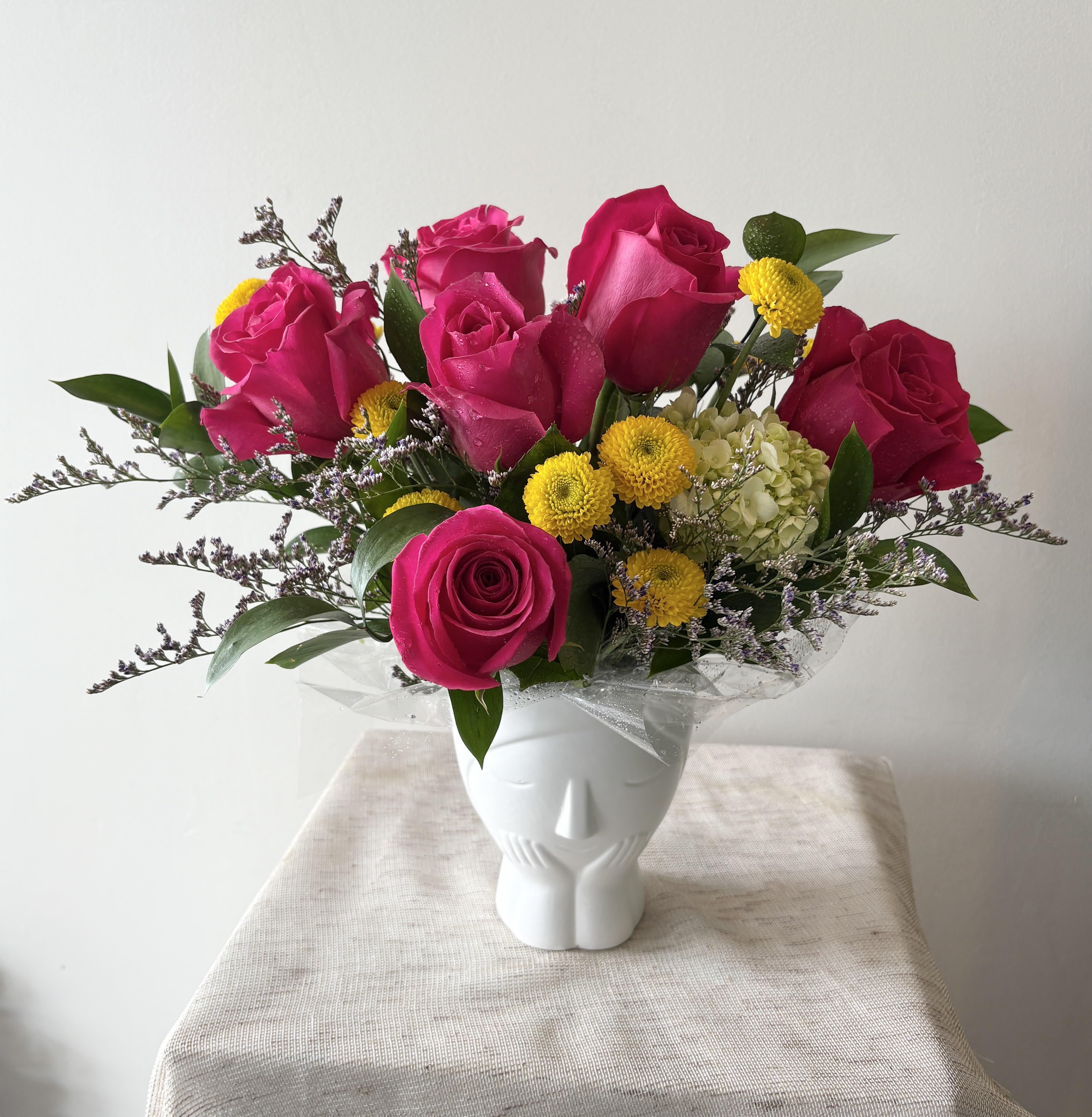Make Me Blush Bouquet - Hot pink roses with some yellow and purple fillers in a white CUTE ceramic pot.  Standard: 6 hot pink roses Deluxe: 8 hot pink roses Premium: 10 hot pink roses