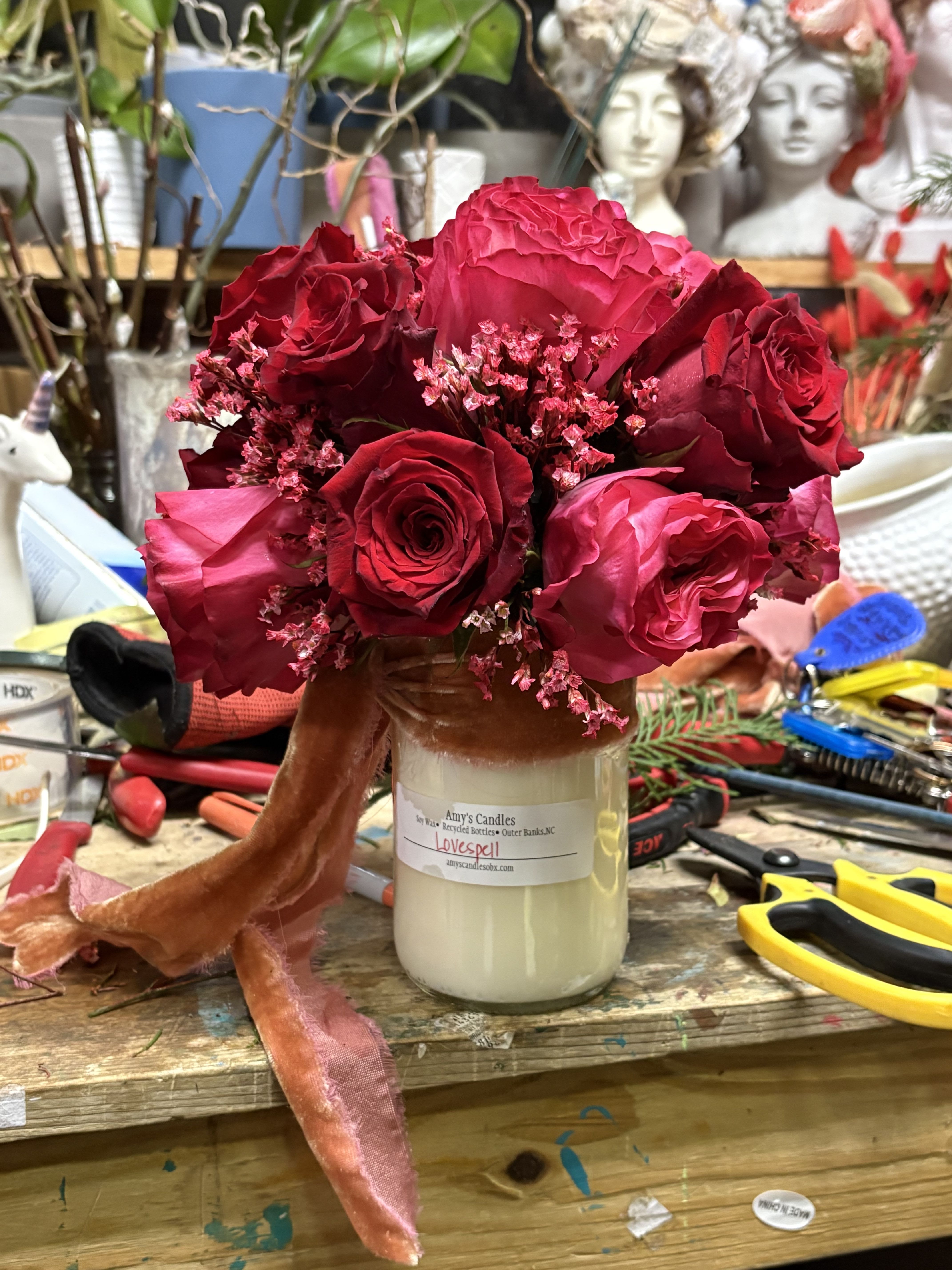 Love Potion - A lush floral arrangement atop a softly glowing candle, tied with velvet ribbon and romantic blooms. This piece brings warmth, comfort, and quiet love to any space—perfect for gifting or creating a cozy, heartfelt moment. Candle made by local artisan Amy’s Candles. Florals may vary with the season.