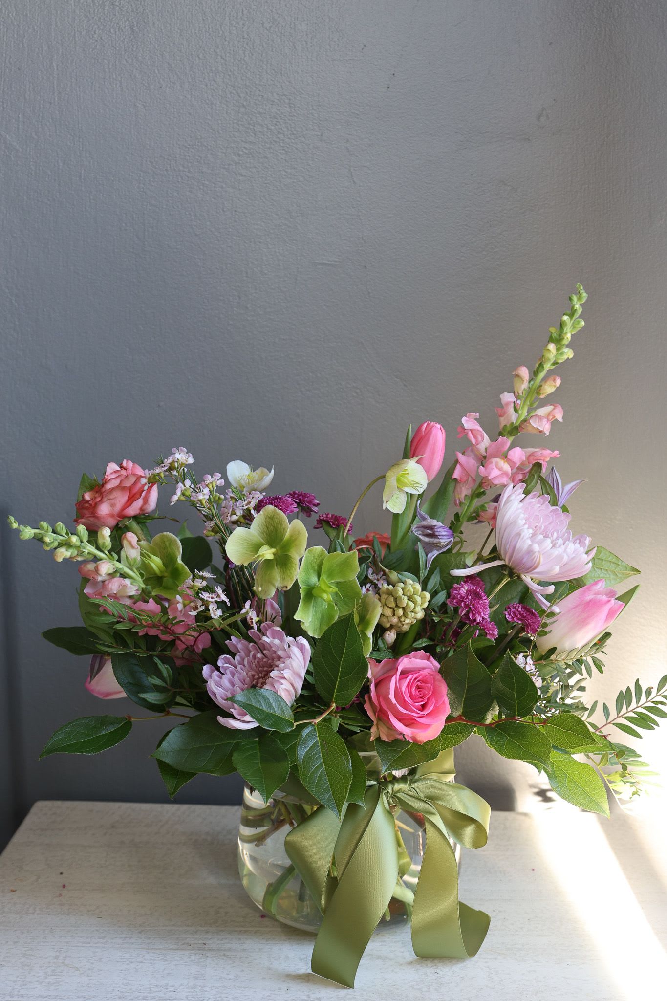 "In Full Bloom" Seasonal Designers Choice - A mix of seasonal florals mixed with lemon leaf and any fun surprises blooms we find throughout the week, will include premium flowers and have a more loose garden appearance  arranged in a 6" clear glass rounded cylinder vase  flowers in photo may vary, and filled to value