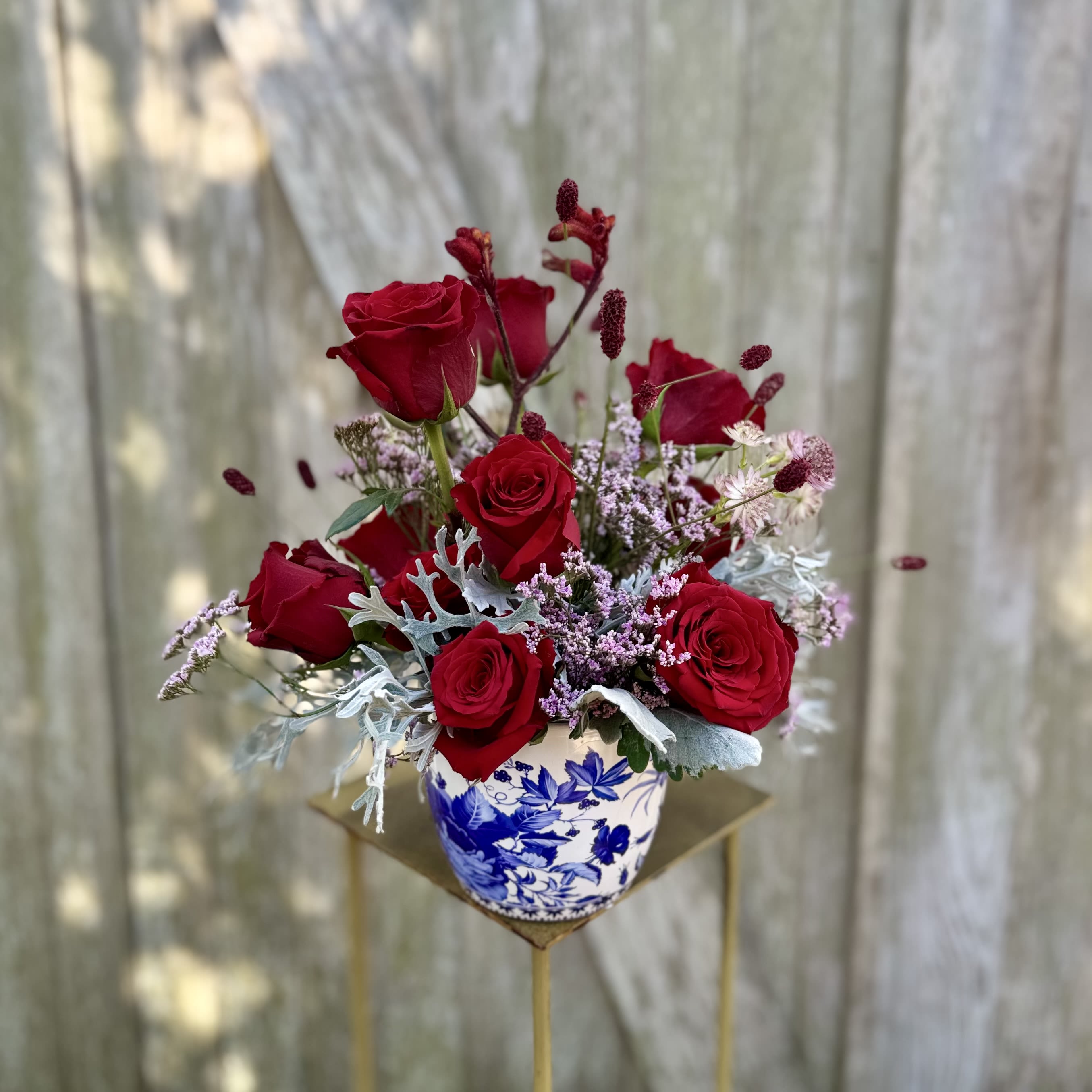 The Heirloom - A timeless arrangement of rich red roses and seasonal accent florals, thoughtfully designed in a blue-and-white ceramic vase. Romantic and refined, this piece makes a striking statement for any occasion. Florals and textures vary based on seasonal availability.