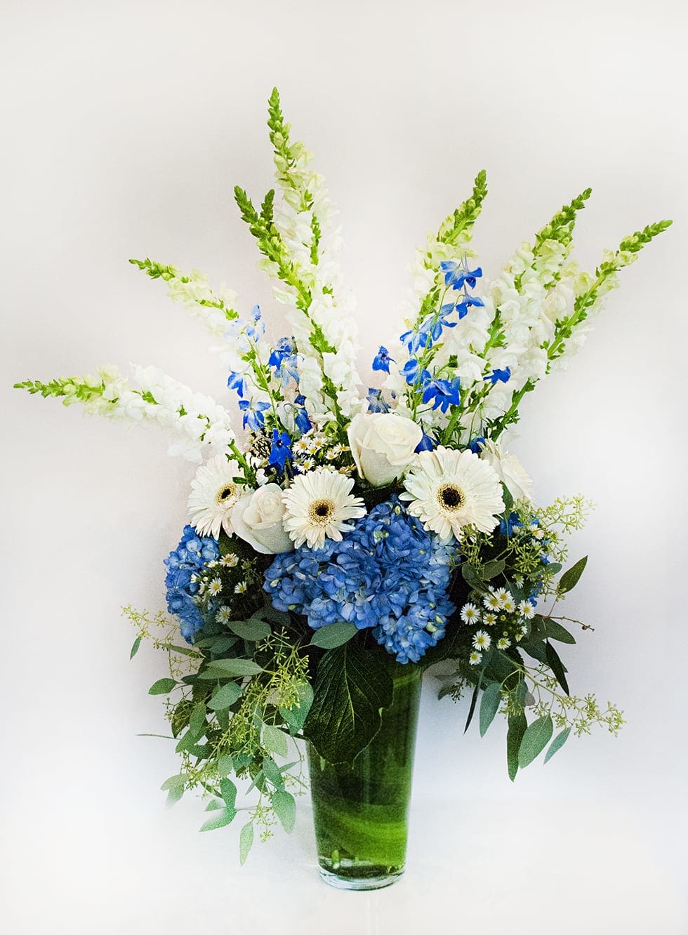 Blue and White  - gerbera daisy's, roses,  delphiniums, snapdragons, hydrangea and greens arranged tall in a clear glass vase with a leaf wrapped to hide stems. 