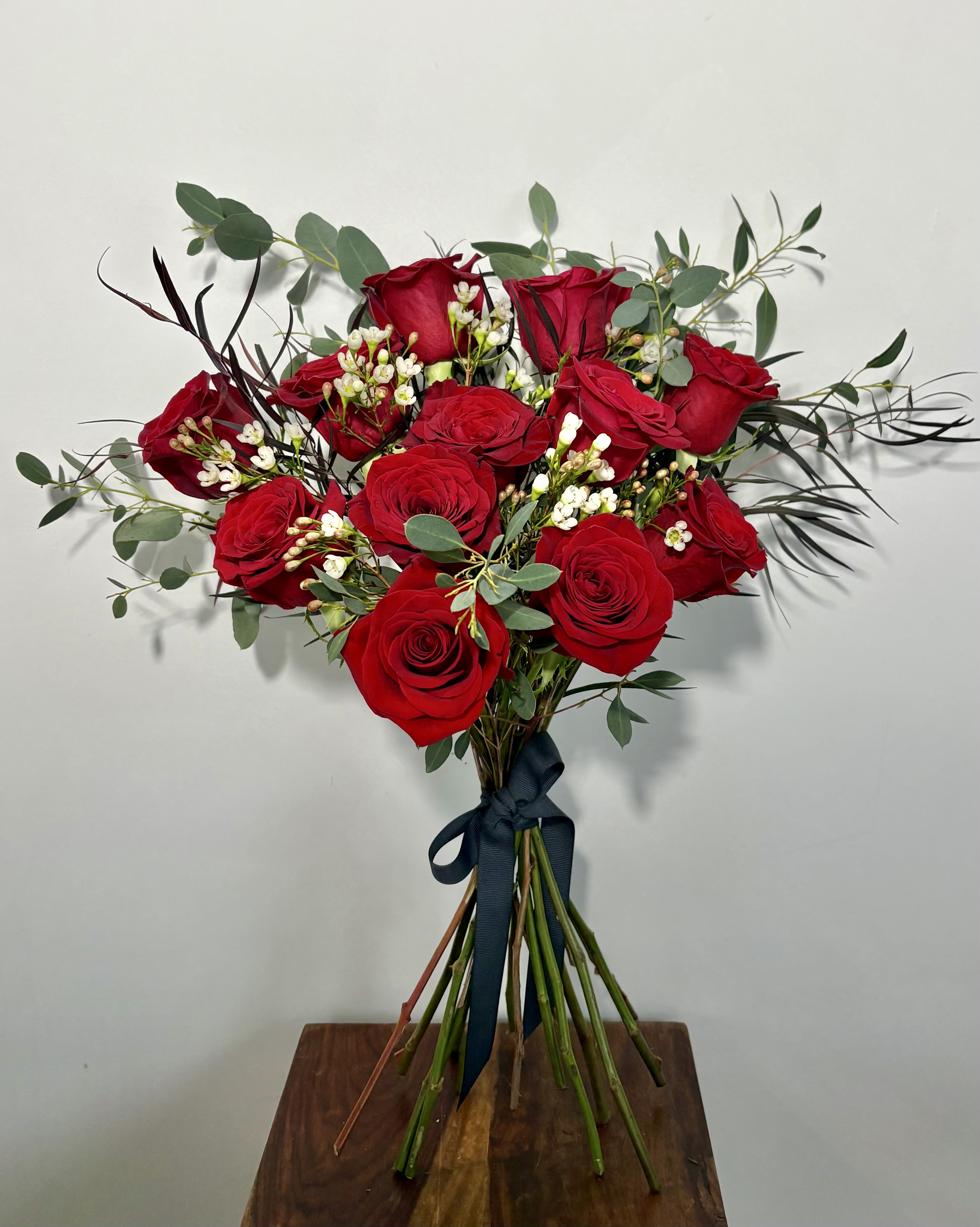A Dozen Classic Red Roses - Timeless and unmistakably romantic, our Classic Dozen Red Roses is the ultimate expression of love. Handtied, featuring twelve velvety roses, thoughtfully accented with airy Wax Flower, touches of Gunni Eucalyptus, and textured Agonis grass, this arrangement is both elegant and lush. Designed as the perfect gift for your partner—or a stunning choice for Valentine’s Day—when only the classic symbol of love will do. + $10 to arrange in glass vase 